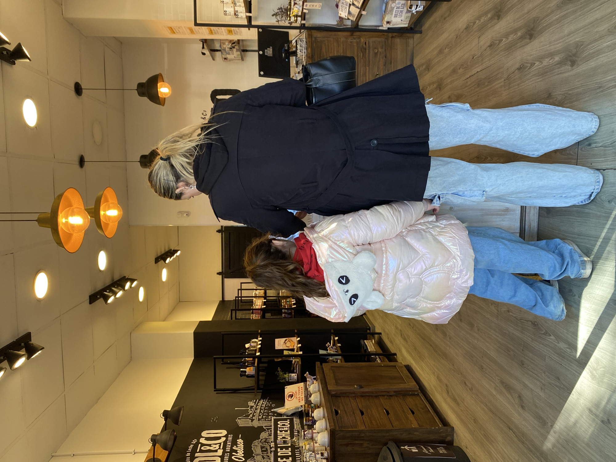Two females are in a retail store that has the signage “DL&CO”. The taller female wears a black hooded coat and light blue jeans. She has a black handbag and is standing next to the shorter female. The shorter female wears a pink quilted jacket with a red hooded shirt. She is wearing blue jeans. The retail store carries a variety of products. There are several lighting fixtures in the store. “Dulce de Leche &Co” is on a shelf. The store has signage that says, “Dulce de Leche &CO, sabros Argentinos Obelisco, nace de leche &co." There is a sign that says “STOP, NO TOQUE”. The floor is wood.
