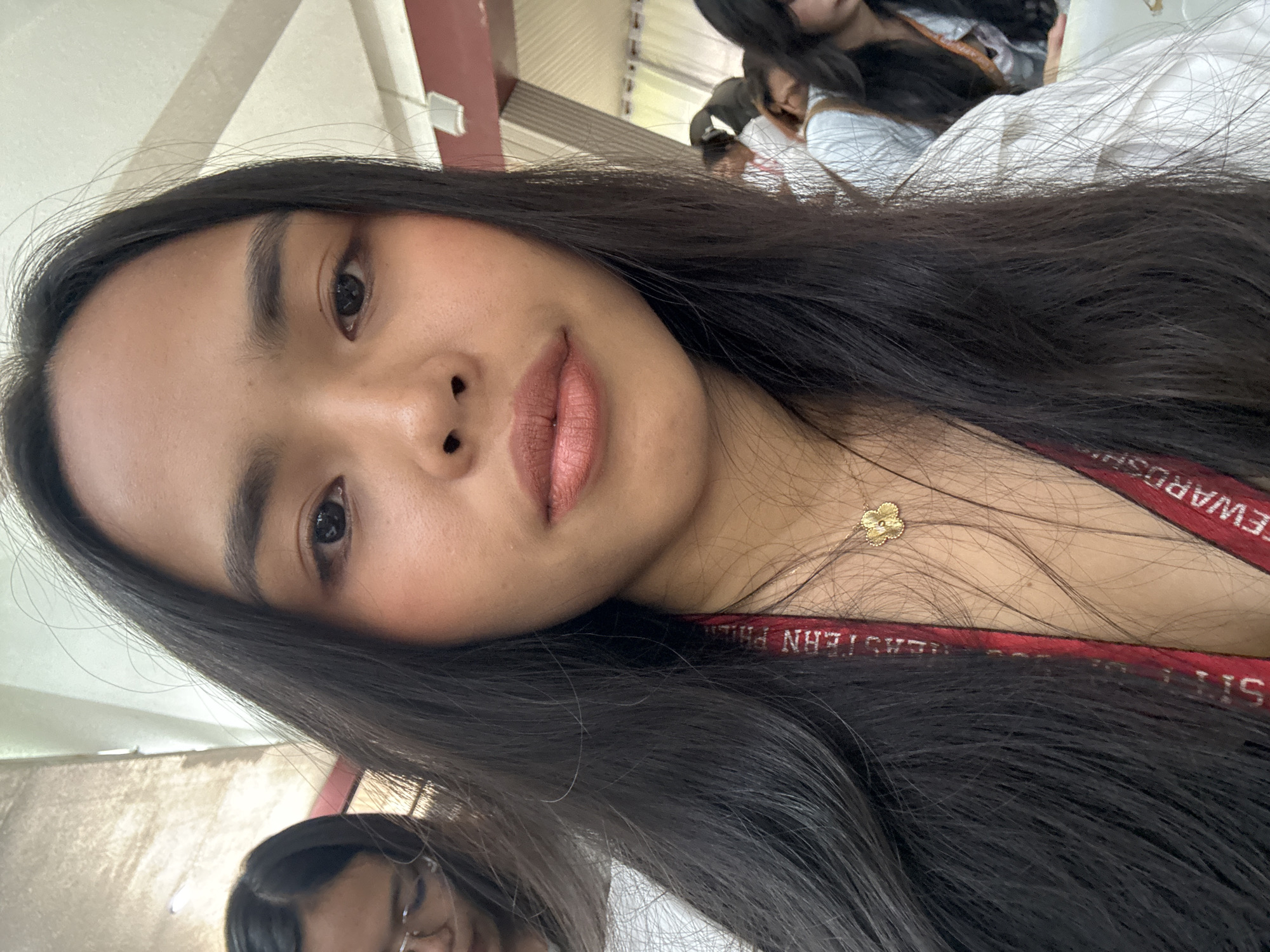 A young woman with long, dark hair takes a selfie. She is wearing a white shirt and a gold flower necklace. She has on light makeup, including blush and lipstick. In the background, there are other people, some of whom are wearing lanyards with text that appears to say "EASTERN P" and "STEWARDSHIP". The setting appears to be indoors, possibly a school or event hall, with a ceiling and a wall visible. This scene is likely located in Davao City, Philippines, given the cultural context and commonality of such events.