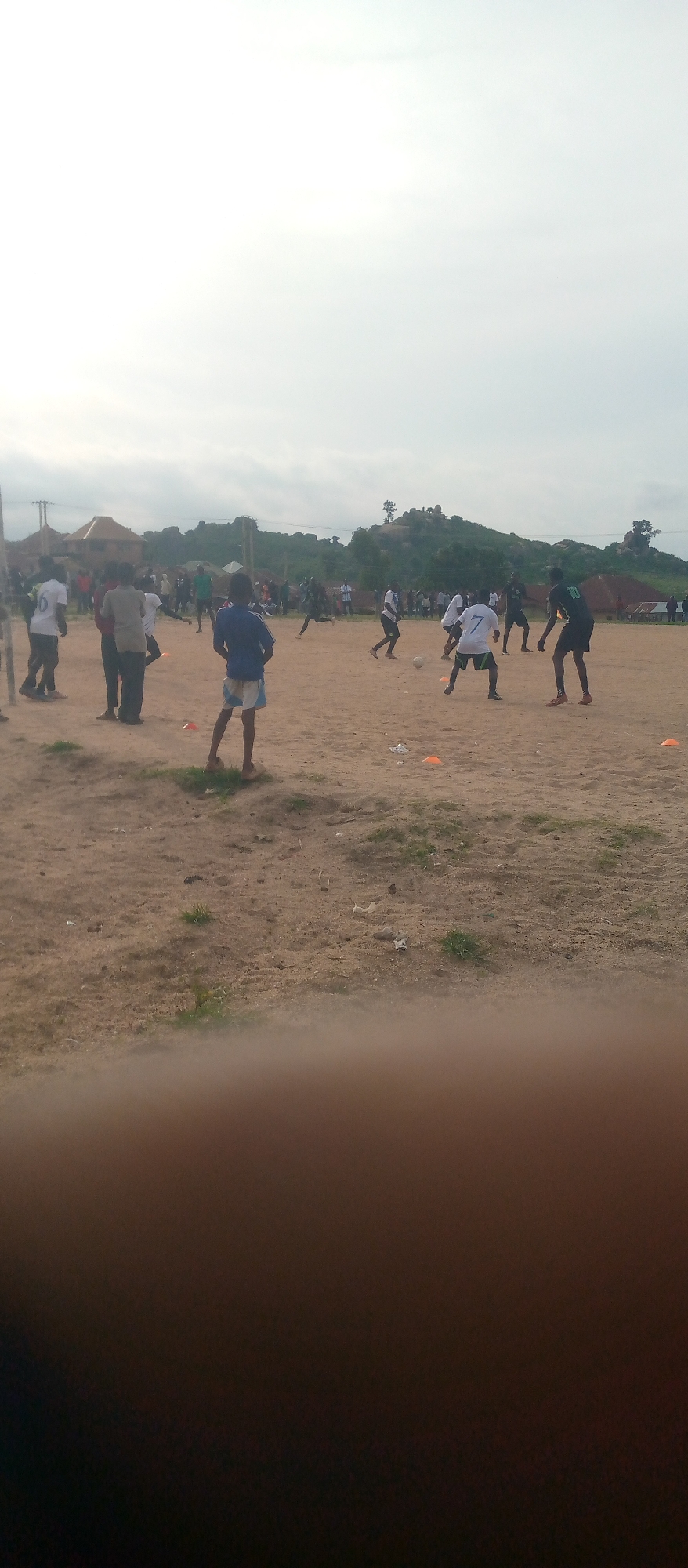In a dusty, open field, a group of individuals, primarily young men, are engaged in a soccer match. Several players in white jerseys and shorts are actively participating in the game, with one player in a dark jersey also in motion. Other figures, including spectators, are visible in the background, observing the match. The environment is characterized by dry, sandy terrain with sparse patches of grass. Hills covered in vegetation form the backdrop, and some rudimentary structures, possibly houses, are discernible amongst the greenery. The sky is overcast with a bright, diffused light, suggesting it might be daytime. Orange cones are scattered across the field, likely used as markers for the game. The scene depicts a casual sporting event in an outdoor, rural or semi-rural setting.