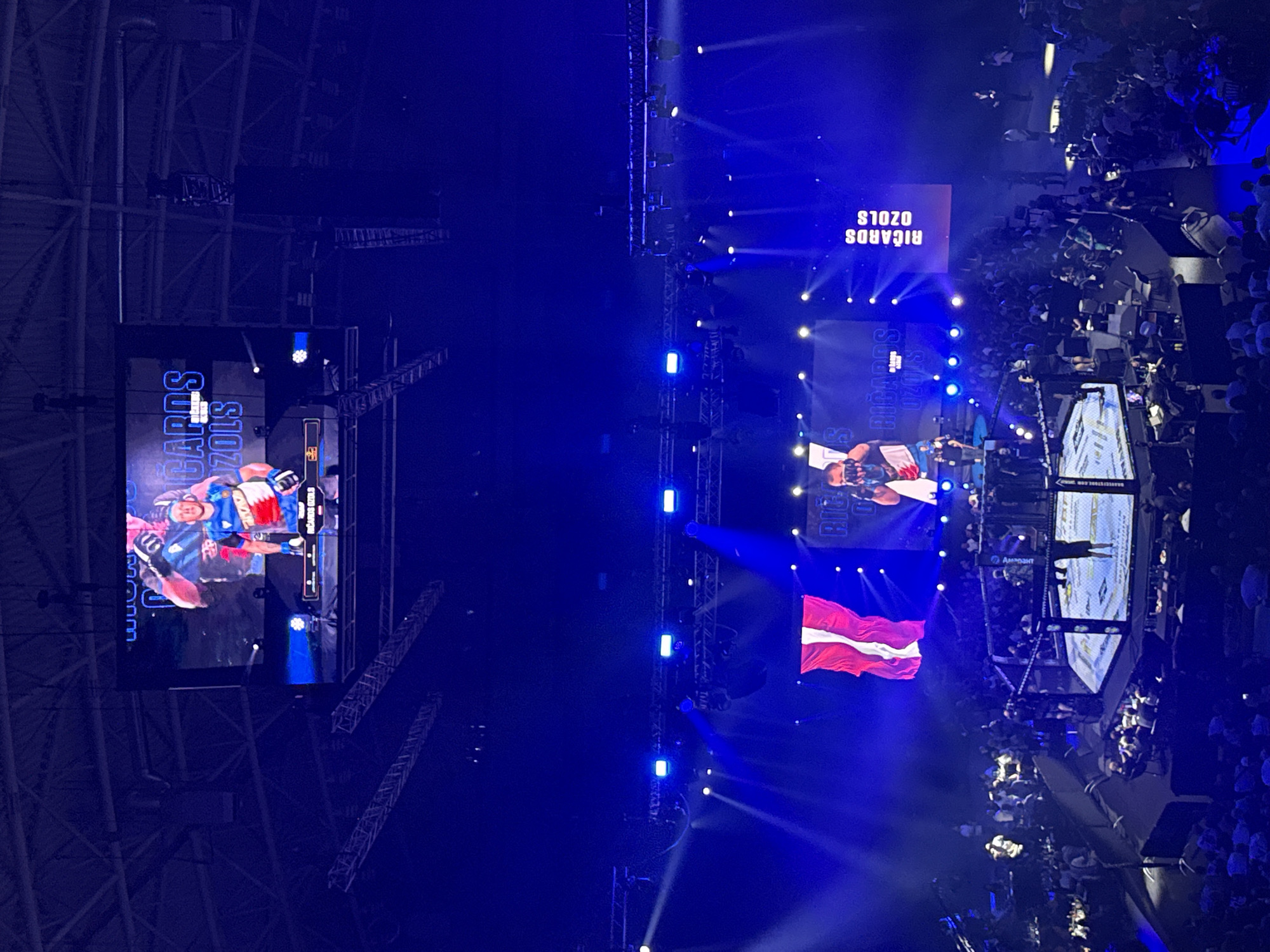The image shows an indoor boxing or MMA event, possibly in Burgas, Bulgaria, given the Latvian flag displayed. A large screen at the top of the arena shows a fighter's name and likeness, possibly "RICARDS OZOLS". Below, on a smaller screen, is a similar display with the same name. The main focus is the octagon-shaped ring where two figures, likely fighters, are present. One fighter, wearing dark shorts, stands in the center of the ring, while another, in light shorts, stands near the ropes. A referee is also visible in the ring. The arena is filled with spectators seated in tiered seating surrounding the ring. The environment is dimly lit, with blue stage lighting illuminating the ring and parts of the crowd. Several cameras are positioned around the ring, suggesting the event is being filmed.