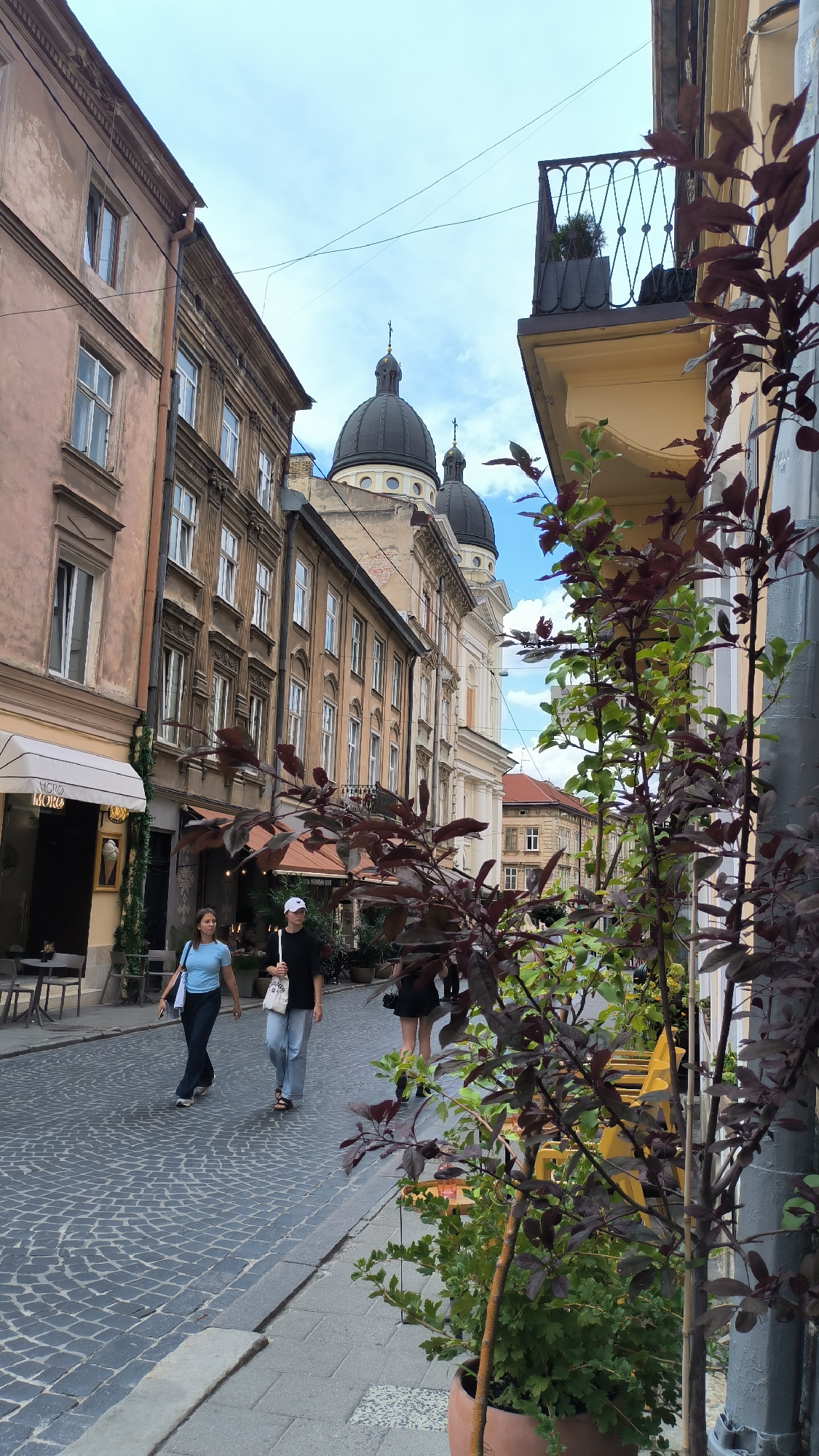 A vertical, eye-level shot captures a European street scene in Lviv, Ukraine, on a cloudy day. The foreground is dominated by lush green and dark red foliage from potted plants, obscuring the lower right portion of the image and extending into the mid-ground. The street itself is paved with cobblestones, with a gray concrete sidewalk running alongside it. Two women are walking towards the camera on the cobblestone street. The woman on the left wears a light blue t-shirt and dark pants, carrying a white tote bag. The woman on the right wears a black t-shirt, light blue jeans, and a white baseball cap, also carrying a white tote bag. Behind them, buildings line both sides of the street. The buildings on the left are older, with weathered, pinkish-brown facades and numerous windows. A sign with the word "MORO" is visible above an awning. On the right side of the street, buildings are a lighter yellow and cream color, featuring balconies with ornate black railings. In the background, two distinctive black domes with crosses rise above the buildings, indicative of a church or cathedral. The sky is a mix of blue and white clouds. The overall impression is a picturesque, urban environment with a blend of historic architecture and greenery.