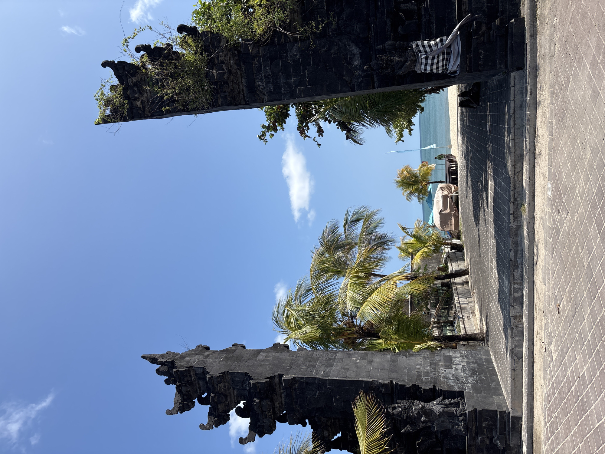 This image depicts a view from a partially obscured Balinese gate in Kuta, Indonesia. The gate is made of dark gray stone and decorated with traditional carvings. Lush green vegetation grows on the top of the gate on the left. Through the gate, the scene opens to a beach, palm trees, and the ocean under a blue sky with scattered clouds. In the distance, there is what looks like a light-colored vehicle, possibly a van. The foreground shows a paved walkway on the right made of rectangular gray tiles. The picture is oriented in portrait.