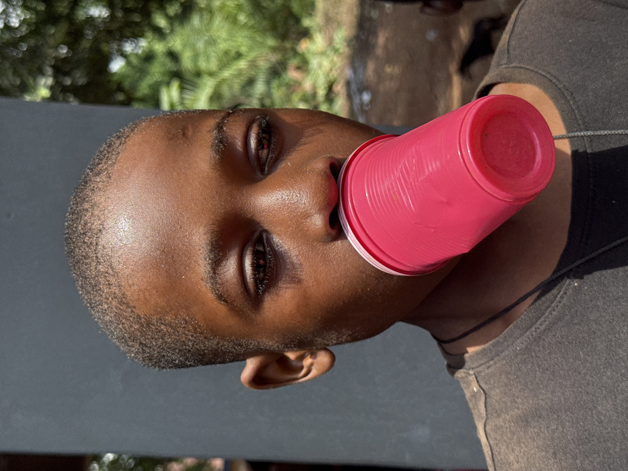 A young, dark-skinned male is seen holding a pink plastic cup to his mouth. He is wearing a dark-colored t-shirt. The location is inferred as Eziudo, Nigeria. A neutral-colored wall is behind the subject, and trees are partially visible in the background. The subject appears to be outdoors in daylight.