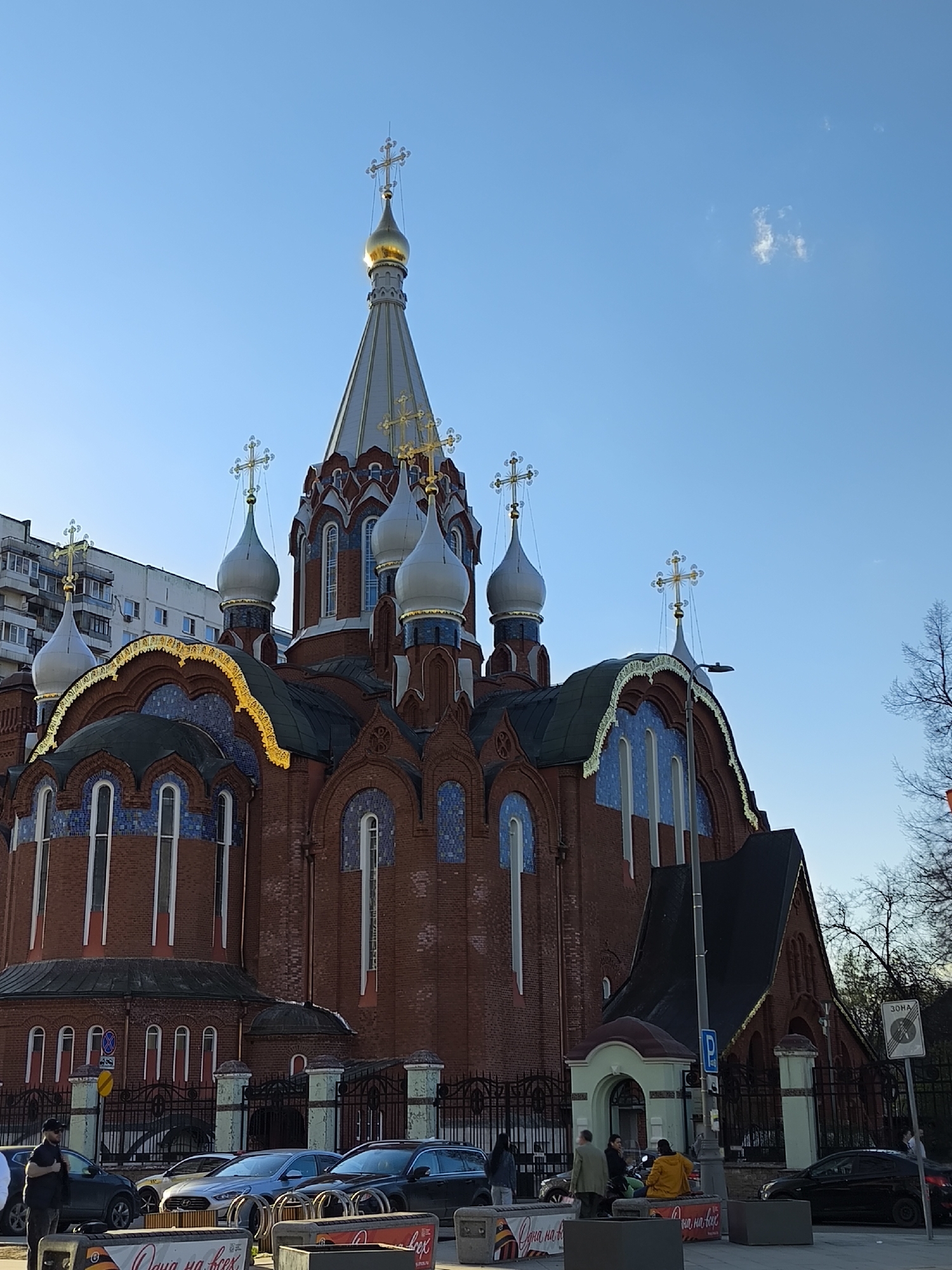 The image depicts a large, ornate Russian Orthodox church with red brick walls, blue and gold accents, and multiple silver domes topped with golden crosses. The main dome is taller and covered in gold. The church is situated in a city environment, with a multi-story apartment building visible in the background. A wrought-iron fence surrounds the church grounds, and several cars are parked nearby. A few people are seen standing or sitting on benches near the entrance of the church. Signage in Russian is present near the benches. The sky is clear and blue. It is likely Moscow, Russia, considering the architectural style of the church and the Russian signage.