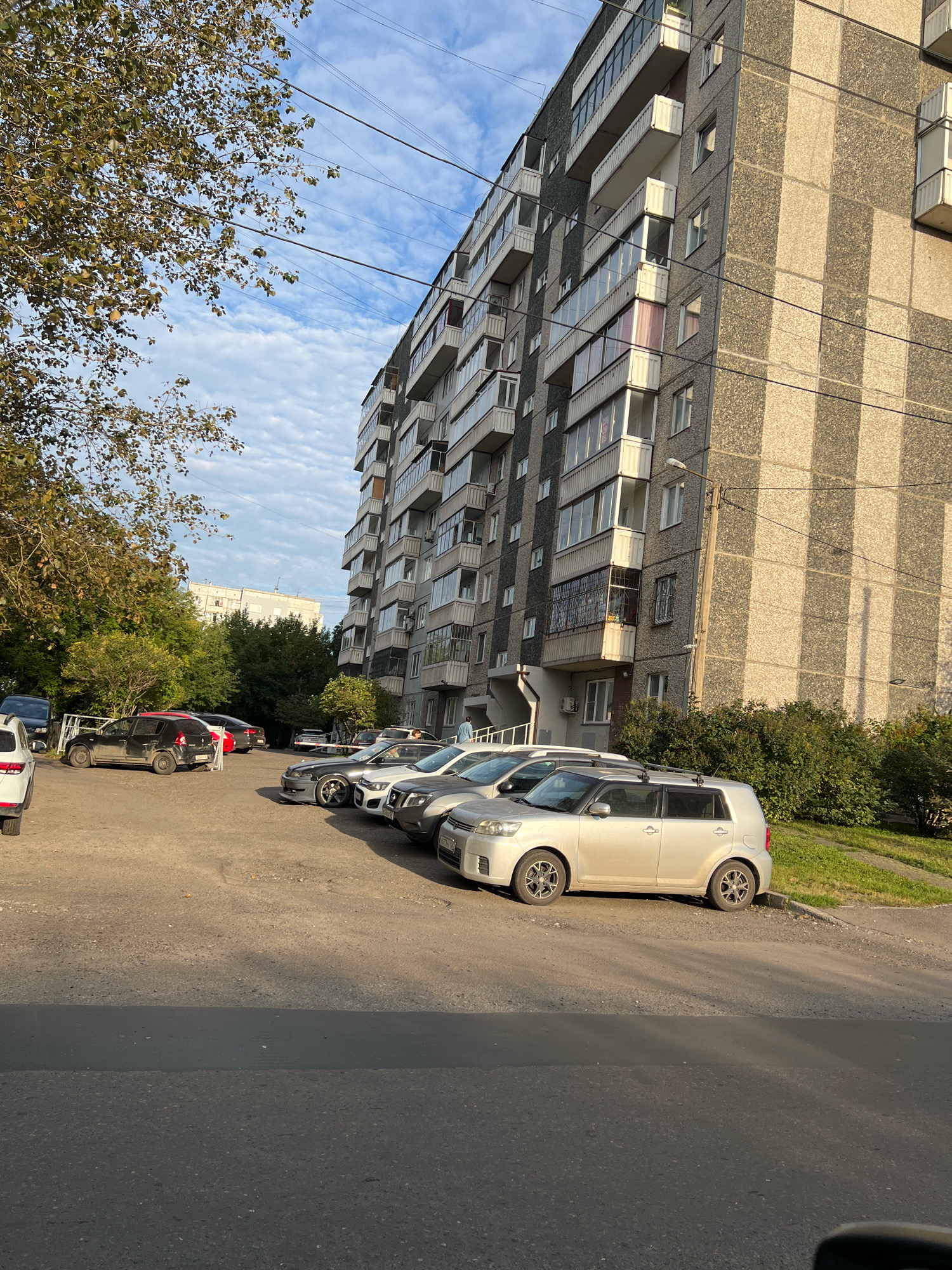 A wide shot captures a residential apartment building in Krasnoyarsk, Russia, on a partly cloudy day. The building, constructed with concrete panels, features multiple balconies and windows. In the foreground, a parking lot is filled with several cars, including a silver hatchback, a black hatchback, and other vehicles. Trees line the left side of the image, with foliage displaying autumn colors. Power lines are visible crisscrossing the sky. The overall scene depicts a typical urban residential area.