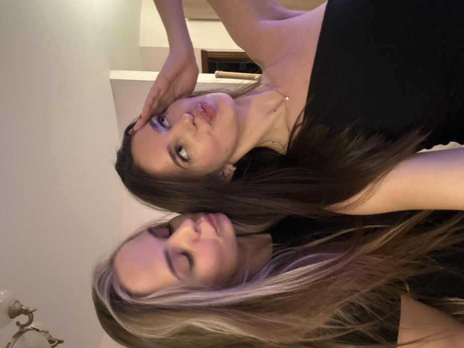 Two young women pose closely for a photo. The woman on top has brown hair, fair skin, and wears a black top. Her hand rests on her forehead. The woman below has blonde-streaked hair and closed eyes. The background includes a white wall, a partially visible window frame, and an ornate light fixture. The image is rotated counter-clockwise.