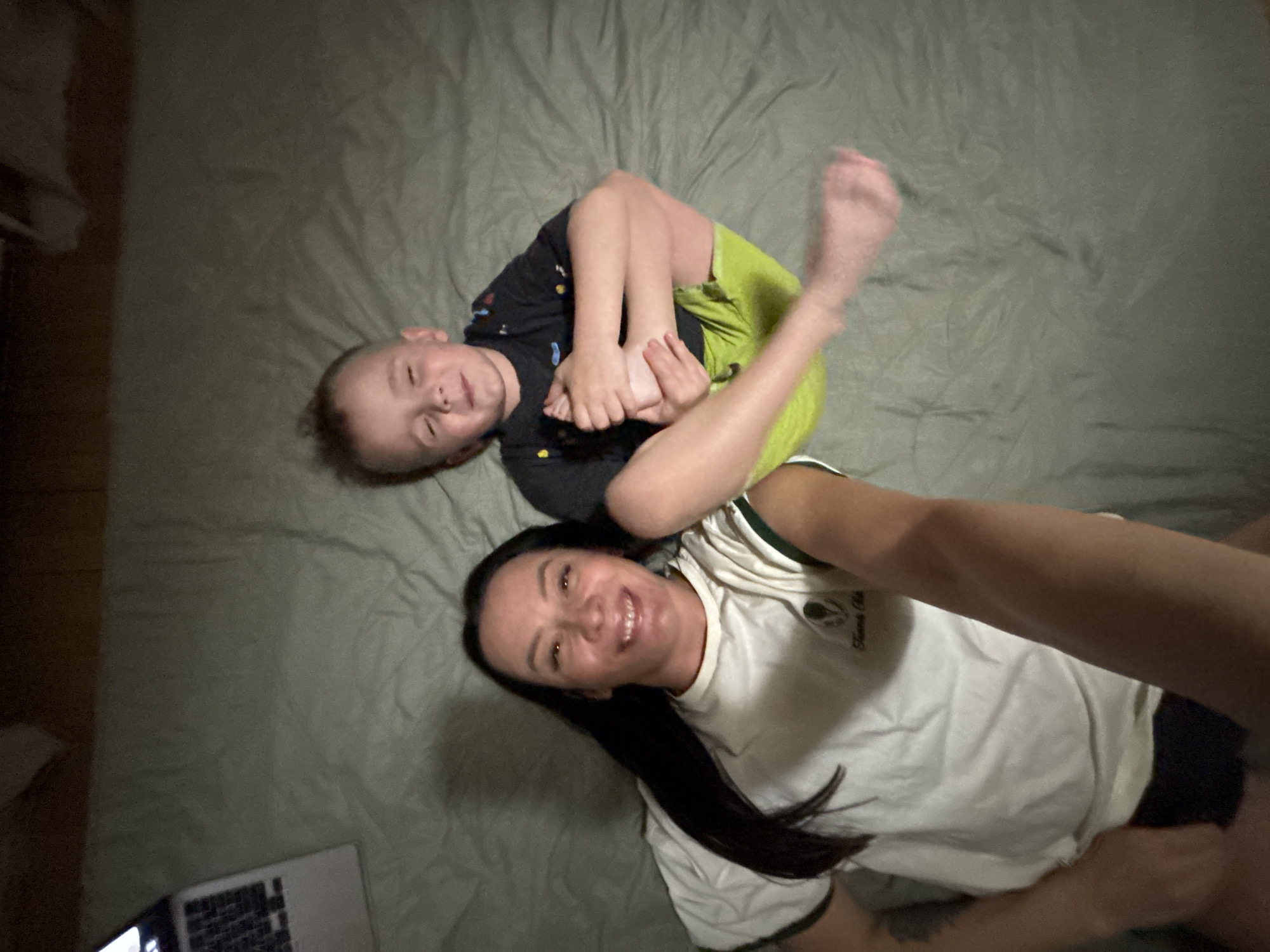A high-angle photograph shows a fair-skinned woman and child lying on a greenish-gray bed. The woman has long dark hair and is wearing a white t-shirt with green trim and black shorts. She is smiling and facing the camera, with her right arm extended towards the camera. The child has fair skin and is wearing a black shirt with colorful designs and bright green shorts. The child is lying on their back with their legs bent, holding their feet. Their right foot is extended upward and is blurry, suggesting motion. A laptop is partially visible in the bottom left corner. The setting appears to be an indoor location. It is not possible to verify the location within the city of Chaikovsky, Russia from the image.