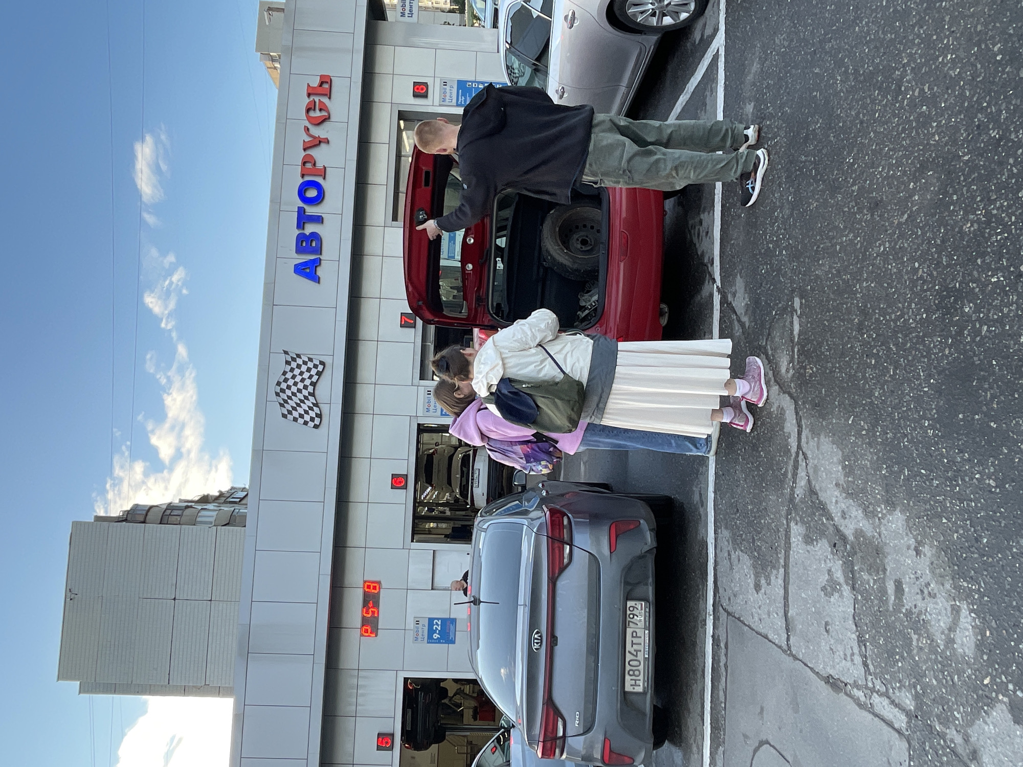 This media file shows an outdoor scene at a car service center, likely in Moscow, Russia, based on the visible text. The setting is a paved lot in front of a building with large signage that reads "АВТОРУСЬ" in blue and red letters. A checkered flag is also visible on the building.
The main subjects are three people. A man in a black hoodie and green pants is standing next to the open trunk of a red car, pointing into it. Two younger individuals, one in a light-colored jacket and long skirt, and another in a purple hoodie, are standing behind the red car, facing away from the camera. A grey Kia sedan is parked to the right of the red car, with its rear facing the viewer.
The time of day appears to be late afternoon or early evening, given the warm lighting and the long shadows cast on the pavement. The weather seems to be clear, with some clouds visible in the bright blue sky. The overall atmosphere suggests a routine car service or interaction at a dealership.
Visible text includes "АВТОРУСЬ," "Mobil 11 Центр," "9-22," "Цена," and the license plate of the Kia, "H804TP 799 RUS." The presence of "Mobil 11 Центр" indicates it's a service or repair facility.