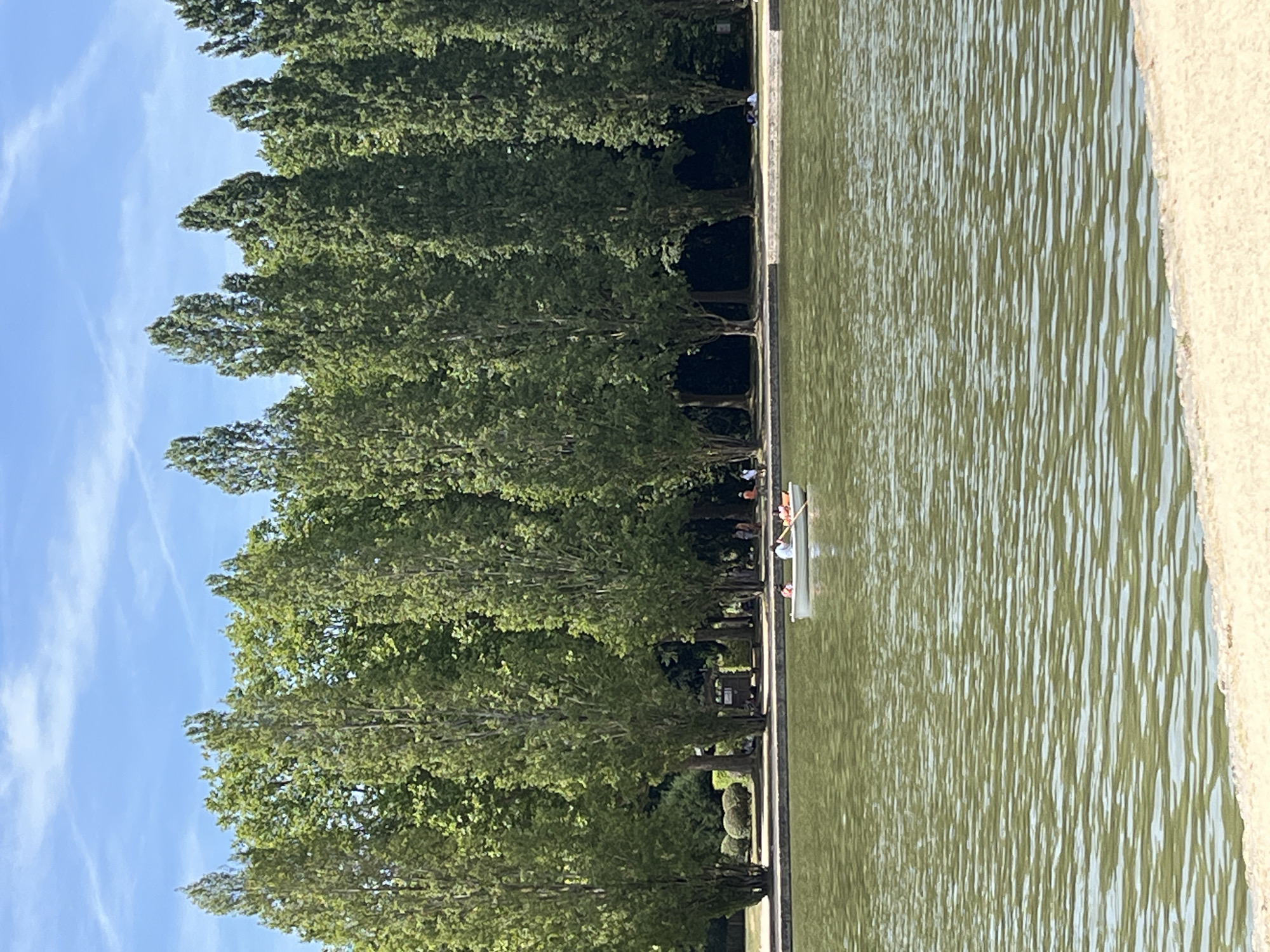 The image captures a serene scene at the Parc de Sceaux, located in Antony, France, on a clear, sunny day. The foreground is dominated by a calm body of water, its surface rippling with a gentle green hue. In the mid-ground, a small white rowboat navigates the water, carrying a few individuals. One person is actively rowing, while others appear to be passengers, enjoying the leisurely activity.
The background of the image is characterized by a dense line of tall, slender trees, their lush green foliage creating a natural backdrop. Behind the trees, a bright blue sky with faint white clouds provides a pleasant contrast. The shoreline is visible, with a paved path running along it, suggesting a well-maintained park environment. Further in the distance, subtle hints of people walking and relaxing on the grass indicate a leisurely atmosphere within the park. The overall impression is one of tranquility and outdoor enjoyment.