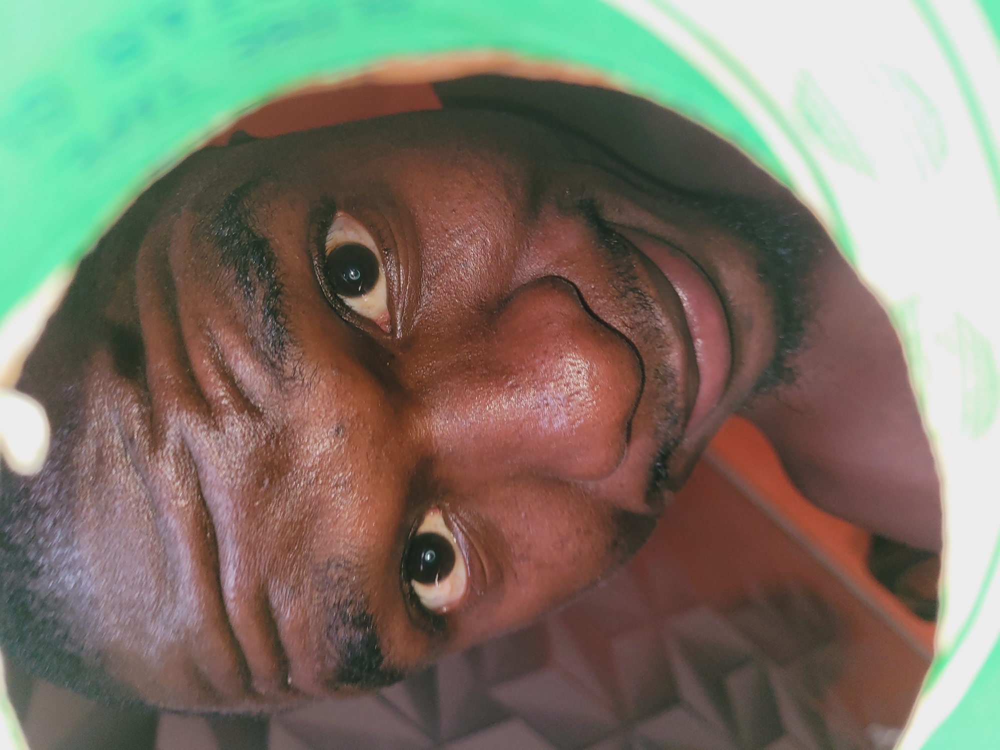 A close-up, low-angle shot captures a young Black man from the chest up, his face emerging from a green circular opening that frames his head. The man's dark skin is dewy with sweat, particularly across his forehead where horizontal wrinkles are prominent. His eyebrows are thick and dark, arching over alert, brown eyes that reflect a subtle glint. The bridge of his nose is broad, and his lips are slightly parted, revealing a hint of teeth and a faint smile, creating a friendly and engaging expression. A light stubble shadows his chin. The green opening appears to be the rim of a container, possibly a bucket or large bottle, with some blurred text visible on its inner surface. The background is indistinct, with a patterned surface in shades of brown and orange suggesting an indoor setting. The lighting is somewhat harsh, creating strong highlights on the man's skin and obscuring finer details in some areas. The overall impression is of a candid and intimate portrait.