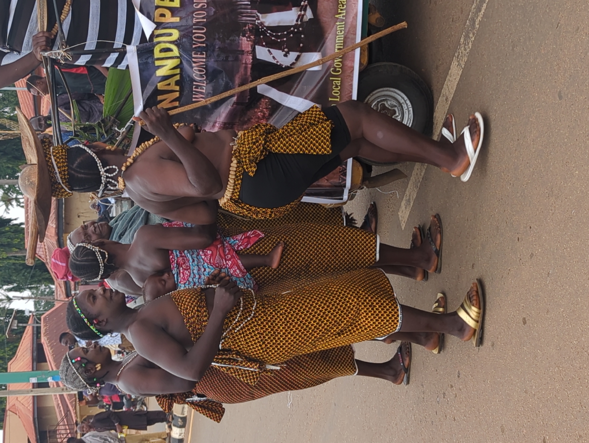 The image depicts a procession of people in Maigizo, Nigeria. Three women in the foreground wear traditional yellow and brown patterned wraps and beaded necklaces. A fourth figure wears a similar wrap with black shorts and a large straw hat. They are all walking on a paved road.
Behind them, a banner reads "NANDU PE... WELCOME YOU TO S..." with "Local Government Area" visible on the banner. A vehicle with a spare tire is partially visible behind the banner. Additional people are visible in the background.
The environment includes buildings and trees.