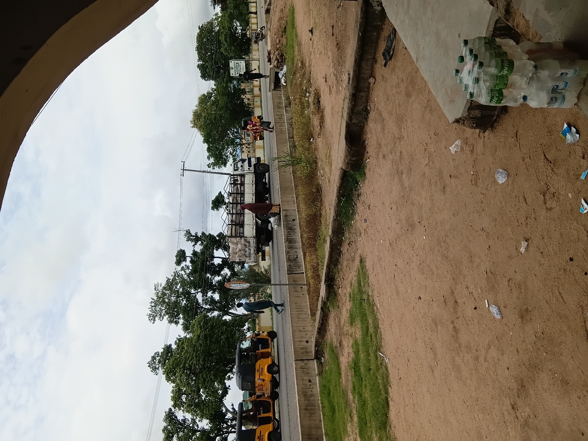 The image depicts a street scene in Jalingo, Nigeria. In the foreground, a dirt and gravel area is visible with scattered debris, including plastic bottles and wrappers. To the right, a stack of bottled drinks, predominantly Sprite, is placed on a concrete structure. A road runs horizontally across the midground, with a concrete barrier separating it from the foreground. Several yellow auto-rickshaws, also known as Keke Napep, are parked on the left side of the road. A white truck carrying sacks is traveling on the road, and a man in a dark outfit is walking away from the truck. Further down the road, another person is walking, and a motorcycle is parked on the right. Trees line the background, with a cloudy sky overhead. A sign with text, likely indicating a location or establishment, is visible on the right side of the road. The overall scene suggests a busy urban environment, with elements of commerce and everyday life.
