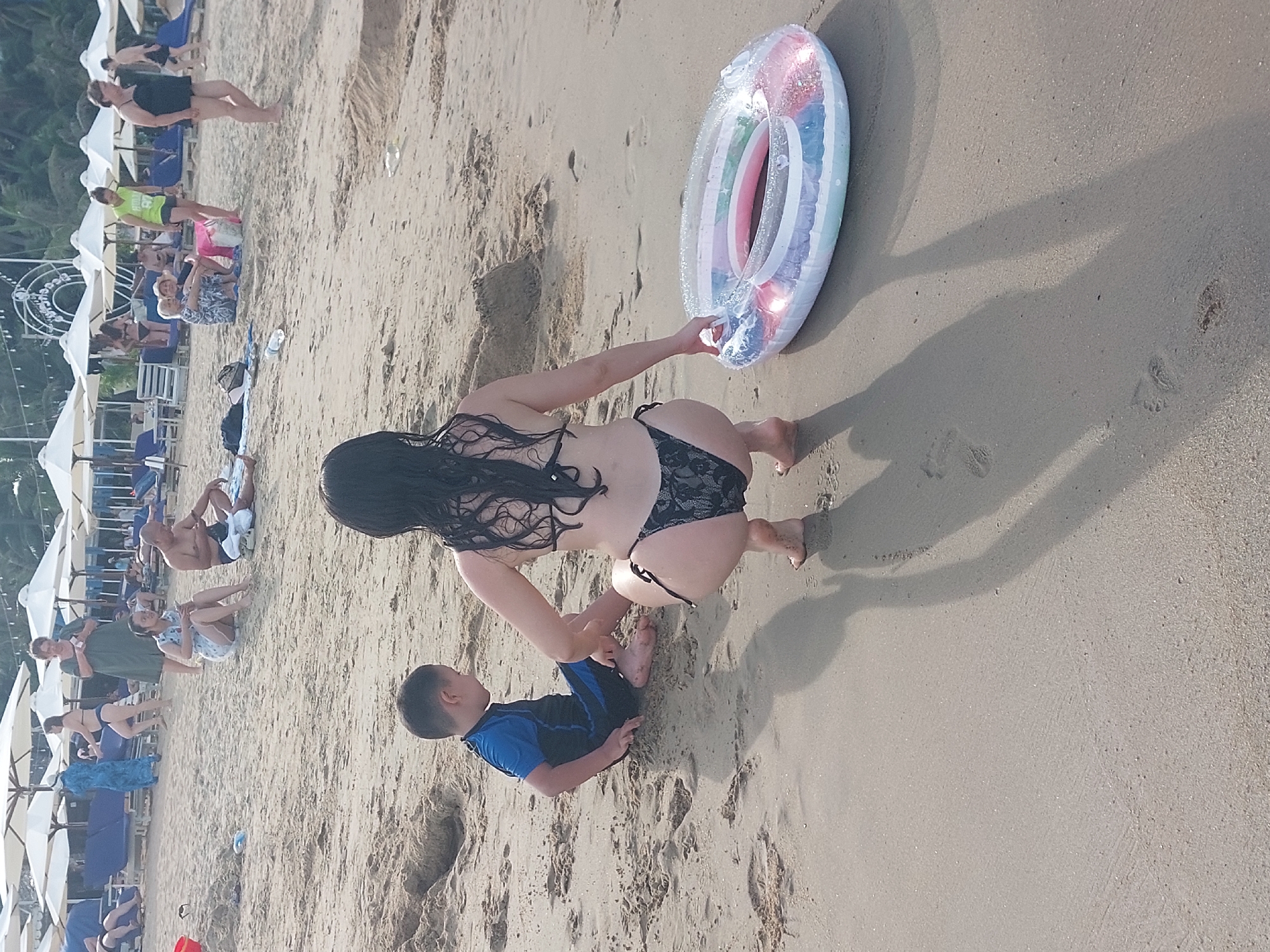 A woman in a black bikini is on a sandy beach, interacting with a young boy also in swimwear. The woman is in a crouched position, reaching towards an inflatable ring. The boy is sitting on the sand in front of her. The beach is populated with other people, many of whom are seated or lying on lounge chairs under white umbrellas. The scene is set during the day, likely late afternoon given the long shadows cast on the sand. The weather appears to be warm and sunny, suitable for a beach outing. The location is identified as Nha Trang, Vietnam, suggesting a tropical or subtropical coastal environment. There are no visible text elements in the image. The overall mood appears relaxed and familial, with a focus on leisure activities by the sea.