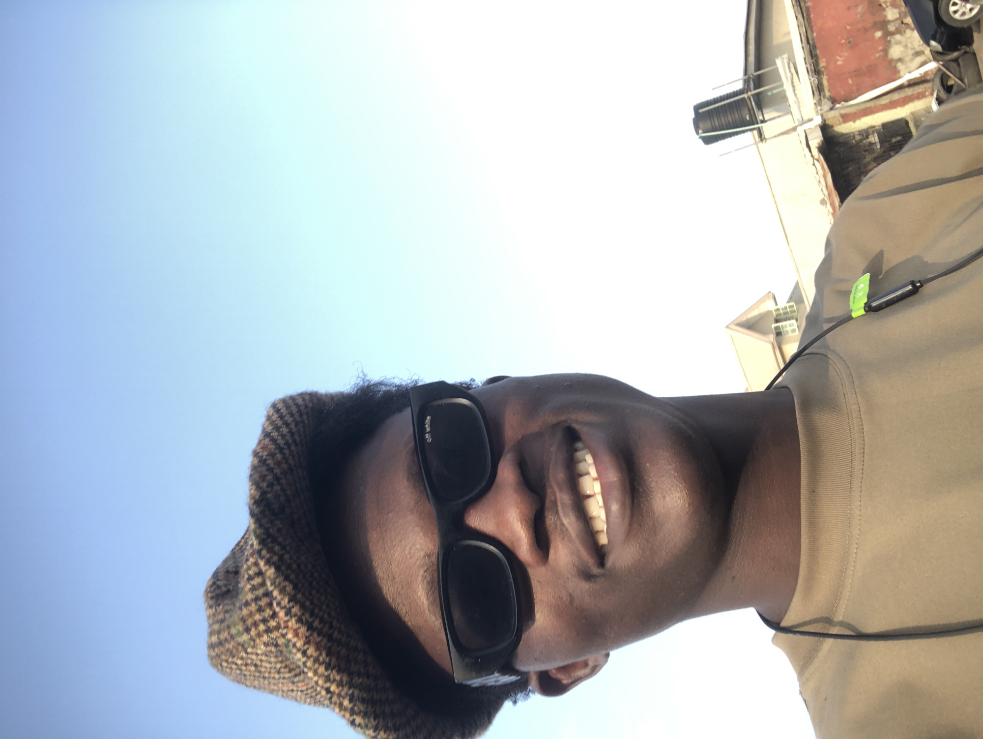 A young Black male is taking a selfie outdoors with a clear blue sky as the background. He is wearing a brown and tan tweed flat cap, black sunglasses with "off white" written on the left lens, and a light brown t-shirt. He has dark curly hair and is smiling, showing his teeth. In the background, there are buildings, including a house with a peaked roof and an industrial-looking structure with a water tank on top. Some tires and a black car are also visible. The scene appears to be in Ado, Nigeria.
