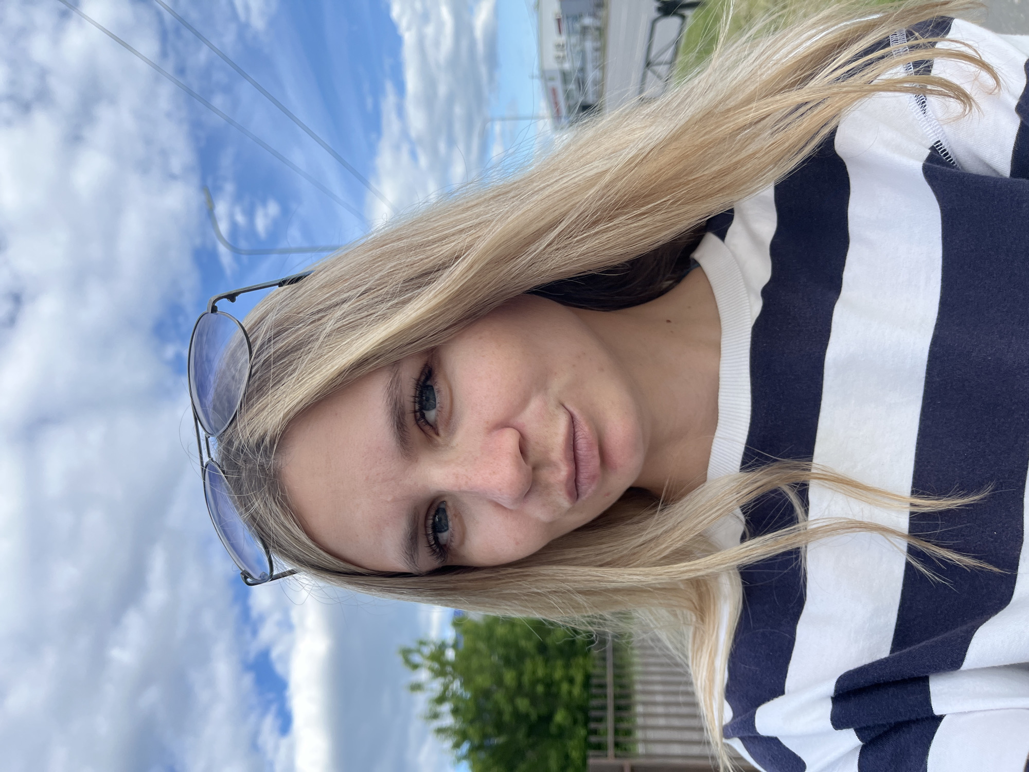 A young adult female is depicted in a close-up selfie taken outdoors, located in the city of Chaikovsky, Russia. She has long, straight, light blonde hair that is parted centrally and extends over her shoulders, with some strands appearing windblown. A pair of aviator-style sunglasses with dark, reflective lenses rests on top of her head, pushed back into her hair. Her blue eyes are directed straight at the camera. Her fair complexion shows scattered freckles across her nose and cheeks. Her lips are distinctly pursed into a pout, and a small, reddish mark is visible on the center-right portion of her lower lip. She is wearing a long-sleeved, crew-neck garment with wide horizontal stripes in dark blue/navy and white.
The background presents a combination of natural and urban features under a partly cloudy blue sky. To the subject's left, a dark brown wooden picket fence runs horizontally, with dense green foliage of trees or bushes visible behind it. To her right, a paved road or street with a visible white dividing line extends into the mid-distance. Further right, a low-rise, light-colored building, possibly commercial, is discernible. Overhead utility lines and a distant streetlamp fixture are also visible against the sky on the right side of the frame. A low, dark metal barrier or fence runs along the roadside in the right foreground. The scene is illuminated by bright natural daylight.