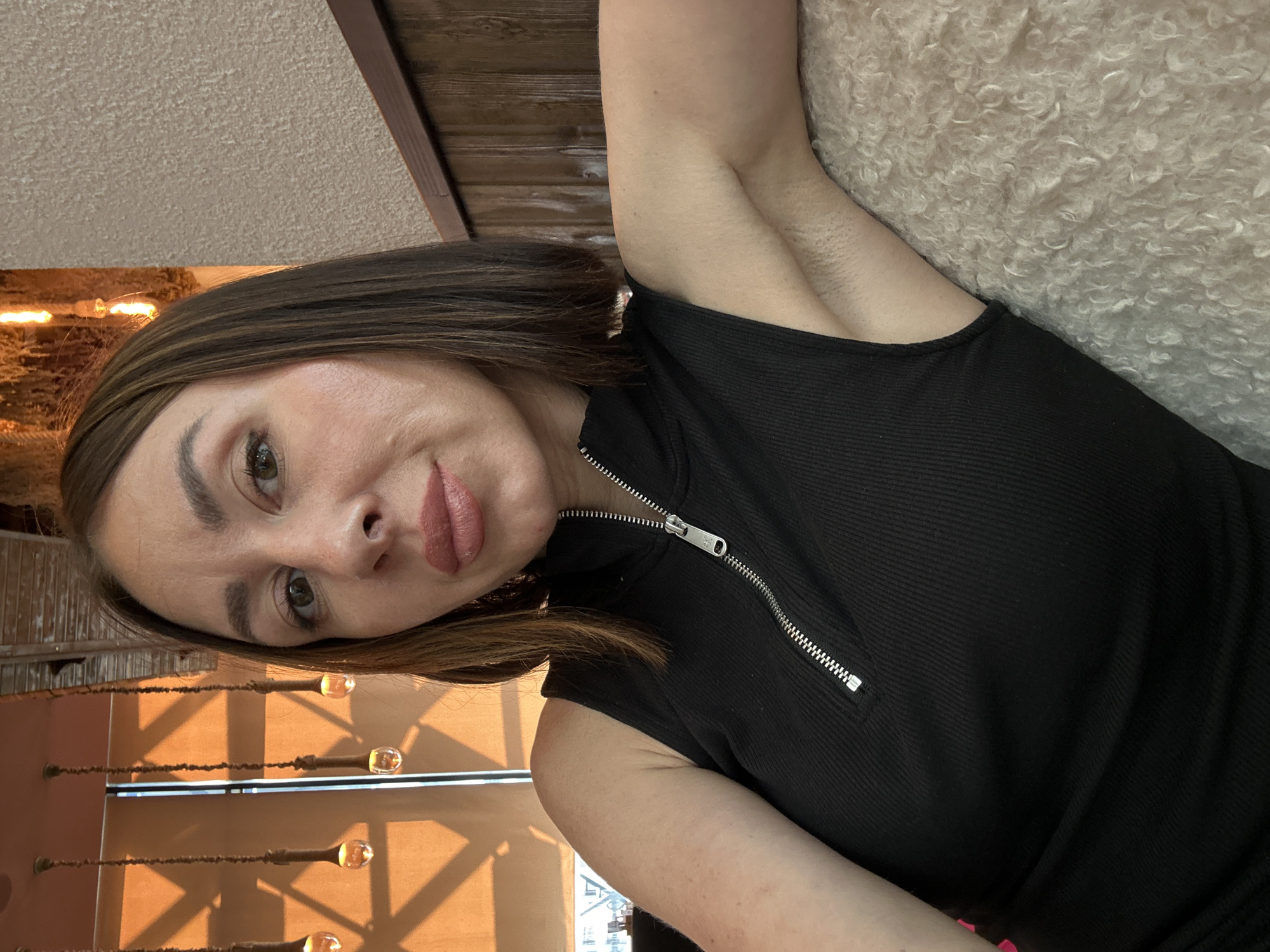 A rotated medium close-up image captures a woman indoors. She has shoulder-length brown hair, brown eyes, and light makeup, including defined eyebrows and pink lipstick. She wears a black, ribbed sleeveless top with a prominent silver zipper down the front, featuring an unidentifiable small symbol on its pull tab. She looks directly at the camera with a neutral, calm expression, hinting at a subtle smile.
The woman appears to be reclining or leaning back against a textured, light-colored, possibly fluffy or sheepskin-like surface. Her left arm, visible on the right side of the frame, shows some fine hair in the armpit area.
The background reveals a warm and inviting indoor setting, potentially a cafe or restaurant in Kazan, Russia. Golden light, possibly late afternoon sun streaming through a window or warm artificial lighting, illuminates the scene, casting soft shadows. In the lower left, several decorative light fixtures with exposed bulbs are visible, contributing to the ambient glow. A wooden beam or structure runs across the upper background, adding to the architectural elements of the space. The overall atmosphere is relaxed and casual.