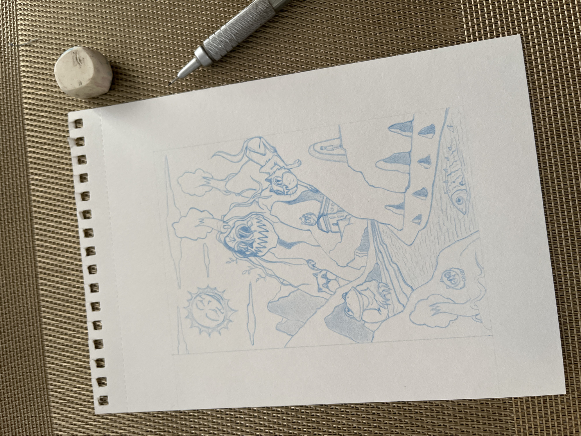 An overhead shot displays an intricate blue line art drawing on a white, spiral-bound notebook page, set against a light brown, woven background. The drawing, oriented sideways on the page, is a detailed and fantastical scene featuring several stylized figures and elements.
Central to the drawing is a creature with wild, flowing hair, sharp teeth, and wide, intense eyes, conveying a sense of ferocity or anguish. Above and to its left, a lithe, feline-like creature with an elongated body and flowing tail appears to be floating amidst stylized clouds or smoke. Other figures include a frog-like creature, a human-like figure wearing a hat, and a small face peering from the side. The scene also incorporates environmental elements such as spiky organic forms, a textured column resembling a tree trunk with an integrated fish, and a sun with a calm, stylized face.
A silver mechanical pencil with a textured grip is positioned near the top right of the paper, and a small, well-used, off-white kneaded eraser rests to the top left. These tools suggest the image captures a moment in the artistic process, likely a detailed sketch phase before inking or coloring. The blue lines are clean and precise, indicating careful execution. The natural daylight illuminating the scene suggests the photograph was taken during the day. There is no visible text on the paper or objects. The setting is a personal workspace, located in Krasnoyarsk, Russia.