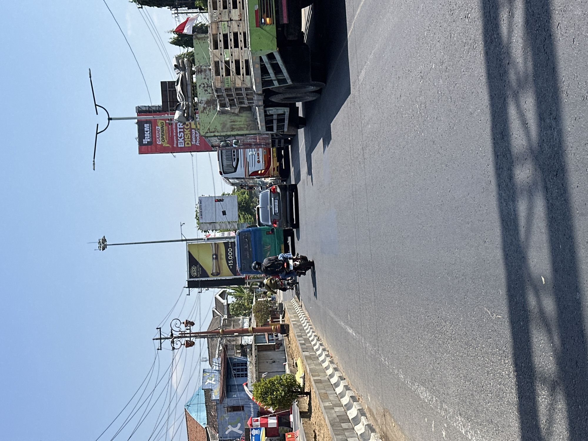 The image depicts a street scene in Bulakrejo, Indonesia, on a sunny day. A line of vehicles, including a green truck loaded with wooden pallets, a blue bus, a black car, and two motorcycles, are stopped or moving slowly on a paved road. One motorcycle rider, wearing a helmet and backpack, is visible in the foreground, while another motorcycle and rider are further ahead.
Along the left side of the road, there are buildings, a decorative lamppost, and electrical wires. A small green tree is planted in a concrete planter. On the right side, a large green truck is prominent, and behind it, a billboard advertises a "GRAND OPENING EKSTR DISKO SEMUA PRODUC." An Indonesian flag is visible on a pole behind the billboard. The sky is clear and blue. The scene suggests traffic congestion.