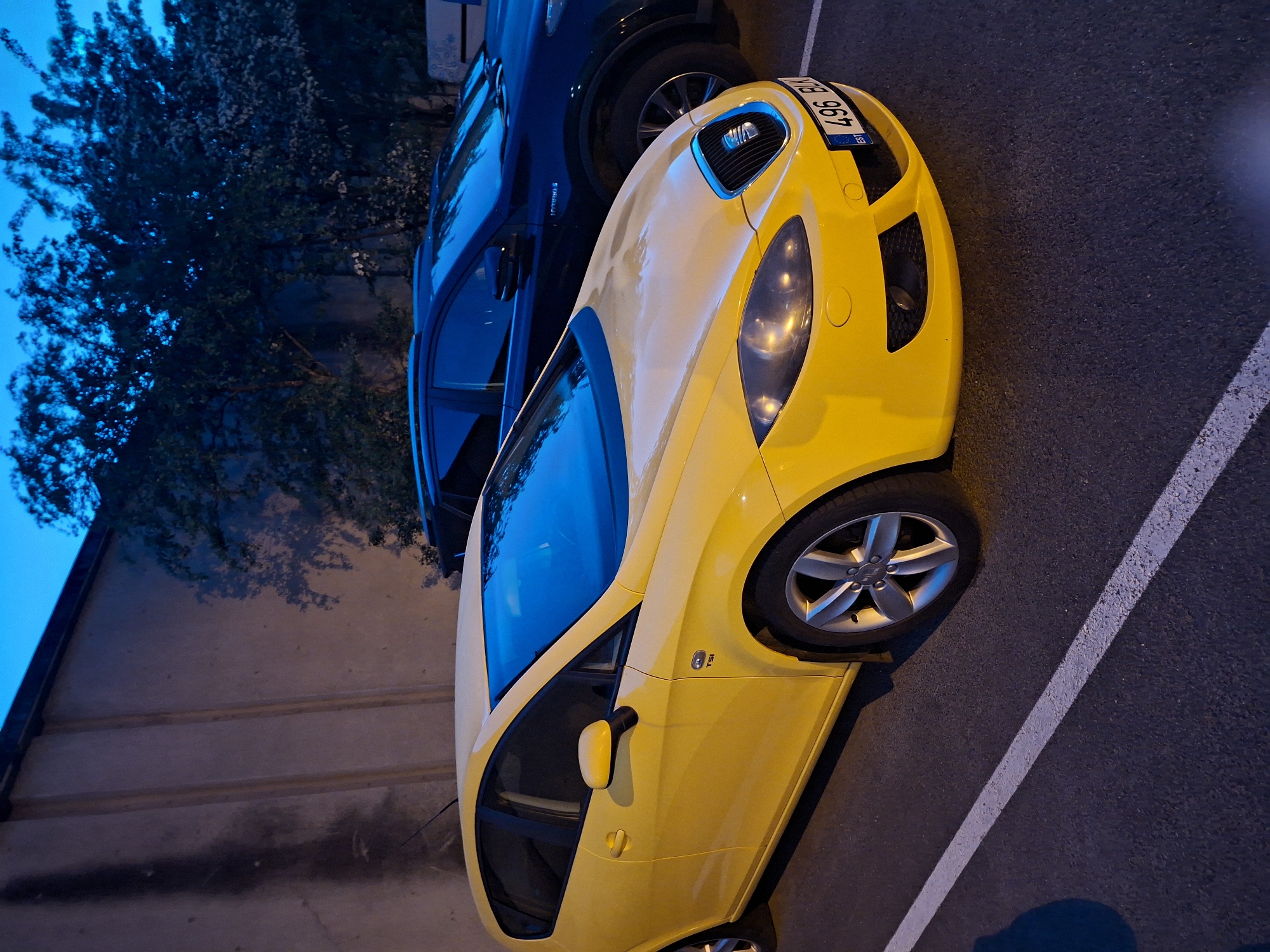 The image depicts an outdoor, urban scene at dusk or night, featuring two parked automobiles in what appears to be a parking lot or roadside in Tallinn, Estonia.
In the foreground, a bright yellow second-generation SEAT Leon (model type 1P, identifiable by its design and "TSI" badge on the fender) is parked facing slightly towards the right. The car is clean, with prominent headlights, a SEAT emblem on its grille, and five-spoke alloy wheels. Its front license plate, "496 BUX," clearly displays the "EST" country code for Estonia, confirming the geographical location within Tallinn as specified.
Behind the yellow SEAT, a darker (likely dark blue or black) SUV or crossover is parked. A "HYBRID S" badge is visible on its side, suggesting it is a hybrid vehicle, possibly a Toyota RAV4.
The ground consists of asphalt pavement, marked with worn but visible white parking lines. To the left and in the background, a tall, light brown or beige concrete wall with distinct vertical paneling is present. A dark, vertical streak, possibly a stain or residue, runs down the left side of this wall. To the right and behind the vehicles, dense trees with dark foliage are visible against a twilight blue sky. No individuals are present in the frame, and no specific actions or interactions are depicted. The scene is illuminated by artificial light sources from the left, highlighting the foreground subjects, while the ambient light from the sky casts a blue hue over the background.
