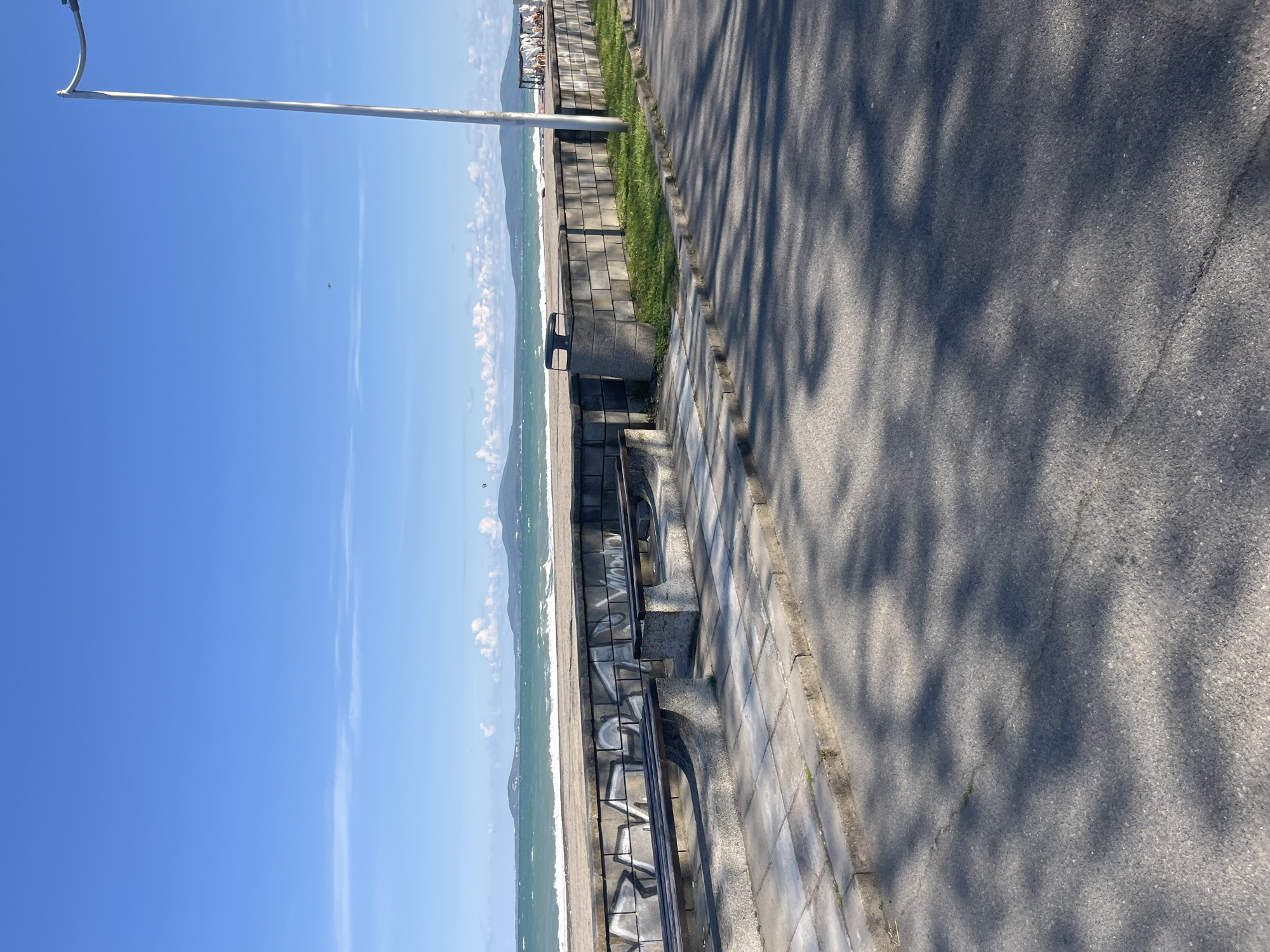 The image depicts a coastal scene in Burgas, Bulgaria. A sidewalk runs parallel to a beach, separated by a low wall with benches, some of which are covered in graffiti. The beach has light-colored sand, and the water shows signs of waves. The sky is blue with a few scattered clouds. A light pole is visible on the left side. The sidewalk is gray asphalt, with tree shadows cast upon it.