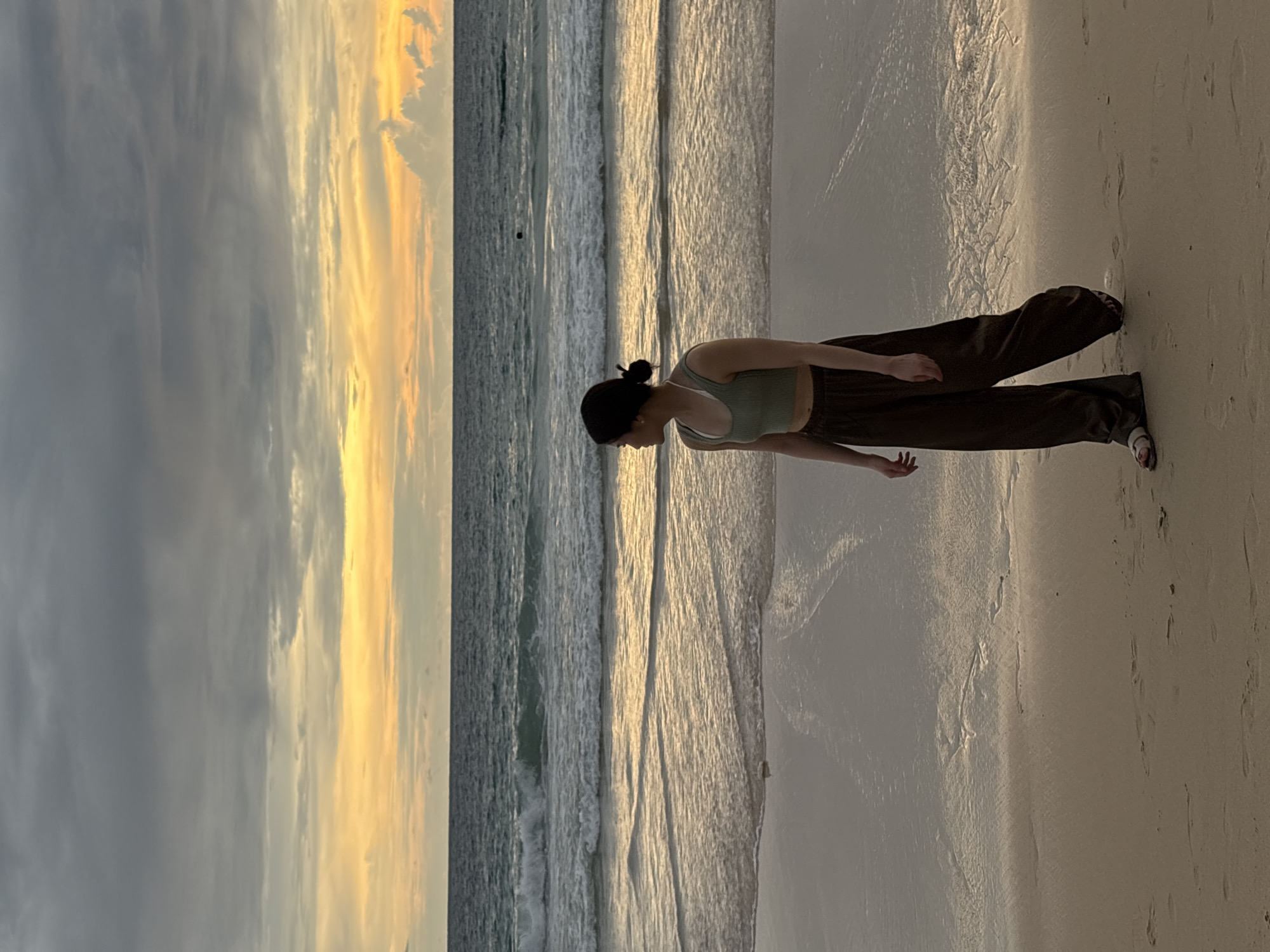 A young woman stands on a sandy beach in Choeng Thale, Thailand, facing the ocean during what appears to be sunset. The sky is a blend of dark gray clouds and bright yellow and orange hues near the horizon, indicating the sun is setting or has recently set. Gentle waves are breaking on the shore, and the wet sand reflects the colors of the sky. The woman is wearing a light green crop top and loose, dark brown pants, with her hair pulled back in a bun. She is looking to her right, away from the camera. Footprints are visible in the sand in the foreground. The ocean stretches out to the horizon, and in the distance, a dark object, possibly a buoy or a person swimming, is visible in the water.