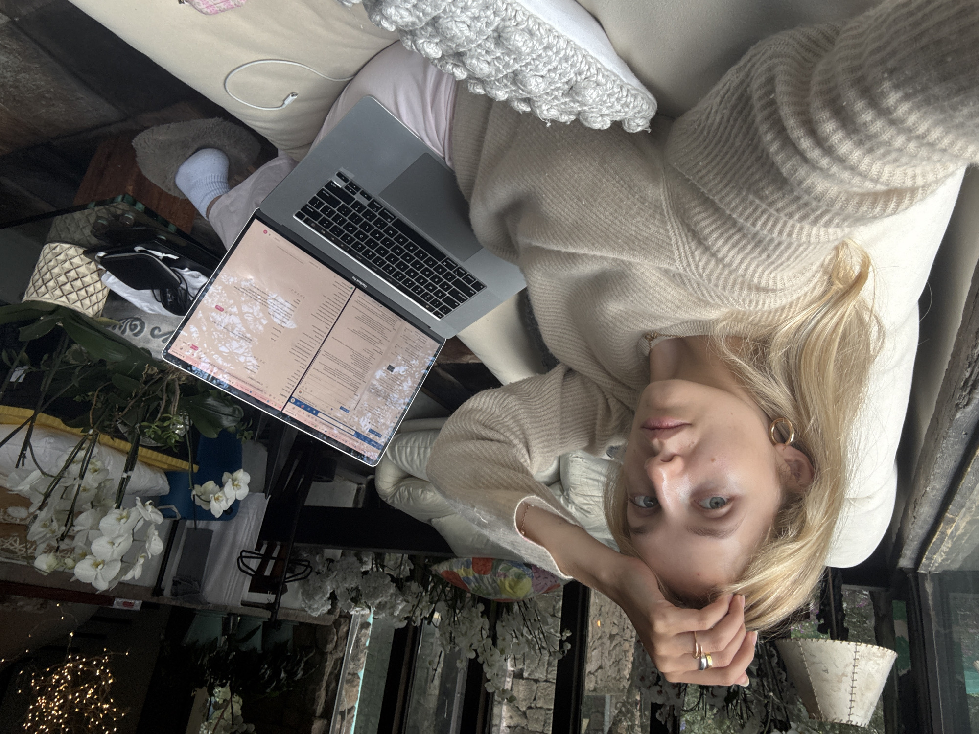 In Mexico City, Mexico, a young woman with blonde hair is pictured indoors, reclining on a cream-colored sofa. She is wearing a beige knitted sweater and pink sweatpants, with white socks on her feet. Her right hand is raised to her forehead, adorned with rings. She is holding a silver MacBook Pro laptop on her lap, displaying text and images on the screen. The background features a large window with a view of vegetation and stone architecture. A white lamp with a textured shade is visible to the left. To the right, an arrangement of white orchids in a pot sits on a glass-topped table, alongside a black bag and other objects. The overall setting appears to be a comfortable, indoor living space.