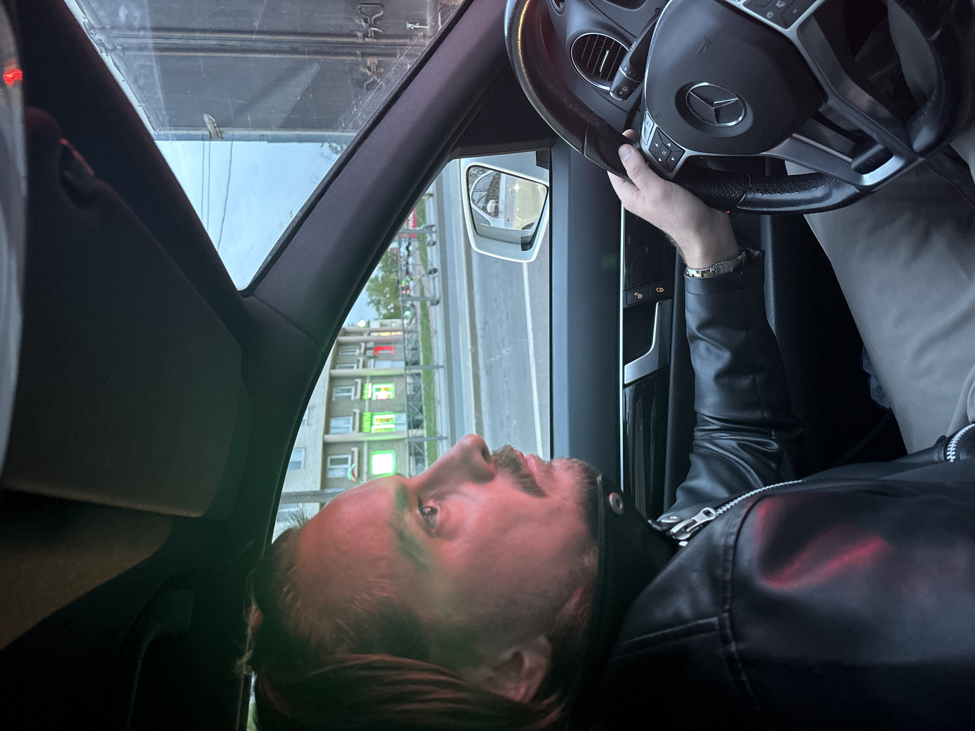 A man is driving a car, his hands on the steering wheel. The car is on a street with buildings visible outside the window, and a side mirror. Based on the style of architecture visible in the background, and the lettering in the illuminated sign, the location is plausibly Saint Petersburg, Russia. The man is wearing a leather jacket and a bracelet. The steering wheel is a Mercedes-Benz steering wheel, based on the logo.