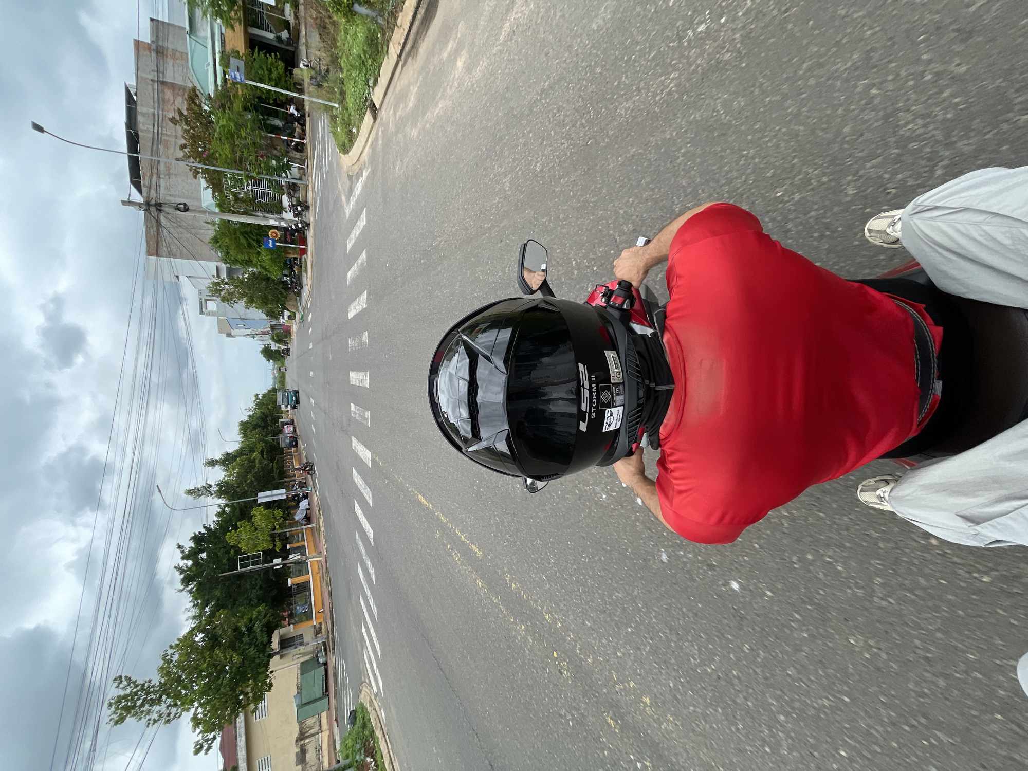 A first-person perspective shot captures a person riding a motorcycle on a two-lane asphalt road, likely in Phu Thuy Ward, Vietnam. The rider, wearing a red t-shirt and light-colored pants, is facing forward, their back to the camera. A black helmet with reflective elements covers their head. The road features white lane markings and a yellow dashed line in the center. On either side of the road, there are sidewalks, vegetation, and various buildings, including a yellow-walled structure on the left and modern, multi-story buildings on the right. The sky is overcast with dramatic clouds. Several other motorcycles are parked along the right side of the road. The overall environment suggests a suburban or developing urban area.