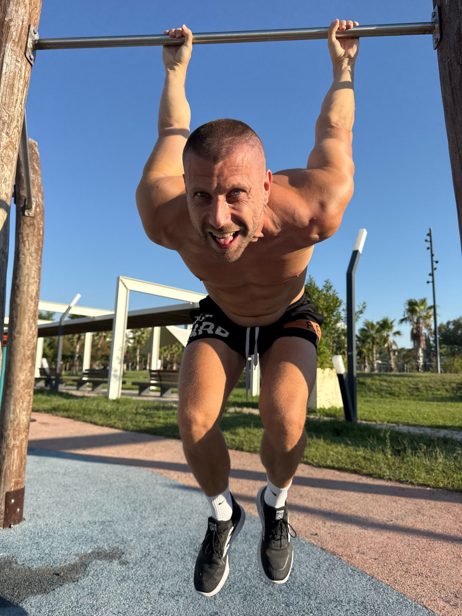 A shirtless man with short hair and a muscular physique is hanging from a pull-up bar outdoors. He is wearing black athletic shorts with an orange stripe and white socks with black athletic shoes. His face is contorted in an expression of exertion, with his tongue sticking out slightly. He is positioned in the foreground, with a park-like setting in the background. This setting includes trees, grass, benches, and a clear blue sky. The ground beneath him is a textured surface with blue and pink sections. The lighting suggests it is daytime. The presence of palm trees and the general landscape are consistent with outdoor public exercise areas found in Antalya, Turkey.