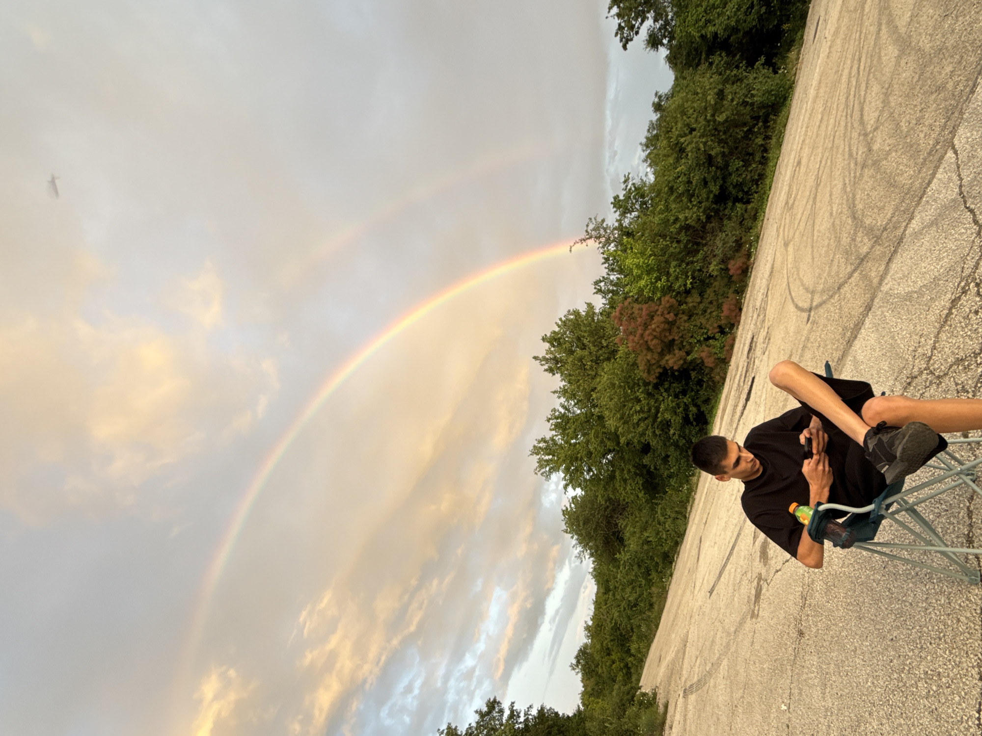 The image displays an outdoor scene featuring a young man seated on a portable chair under a prominent double rainbow, likely in Tuchenitsa, Bulgaria. The man, dressed in a black t-shirt, black shorts, and black athletic shoes, is absorbed in his smartphone, which he holds in both hands. A bottle of a green-yellow sports drink with an orange cap (resembling Gatorade) is visible, hanging from the side of his chair.
He is positioned on a cracked, grey asphalt or concrete surface, which shows faint, dark, circular tire-like marks. Lush green trees, some with reddish-brown foliage, border this open area, providing a natural backdrop.
The sky above features a striking double rainbow, arching from the upper left to the lower right of the frame. The clouds are a mix of light grey and soft, warm-toned patches, suggesting either clearing weather after rain or the soft light of late afternoon or early evening. The presence of the rainbow strongly indicates recent precipitation. In the far upper left corner of the sky, a very faint and indistinct object, possibly a distant bird or aircraft, can be seen. The overall impression is one of peaceful observation, with the natural spectacle of the rainbow providing a serene atmosphere.