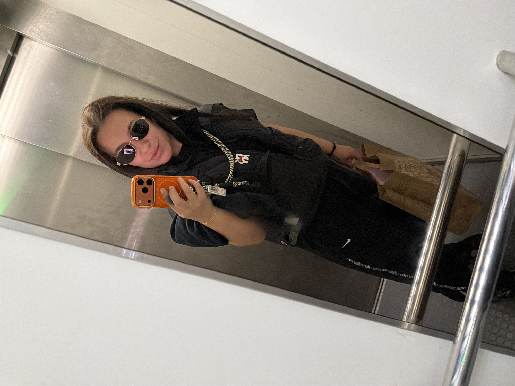 A woman is taking a mirror selfie in what appears to be an elevator. The location is unknown and it is impossible to determine if it is in Burgas, Bulgaria. She is wearing sunglasses, a black vest over a black hoodie, and black athletic pants with a white Nike logo. She is holding an orange phone. There is a brown paper shopping bag next to her. The elevator has stainless steel walls and handrails. The lighting is bright and uniform.