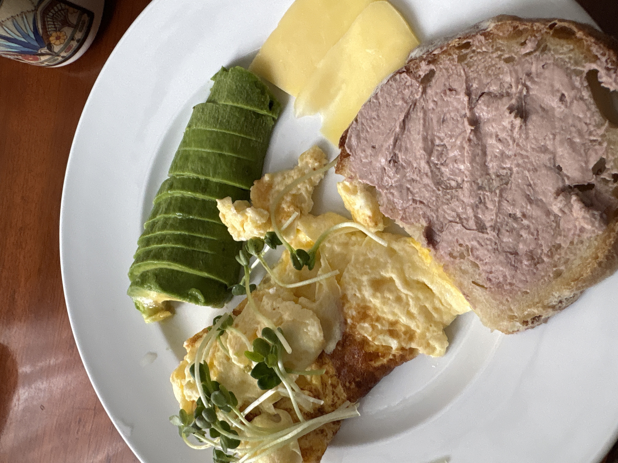 A close-up, overhead shot shows a plate of food on a wooden table. The plate contains scrambled eggs topped with microgreens, sliced avocado, two slices of yellow cheese, and a slice of bread topped with pâté. The food appears to be a breakfast or brunch. There are no people or animals visible in the image. The location is not specified in the image or the provided context, thus it is impossible to determine if it is in Nha Trang Ward, Vietnam.
