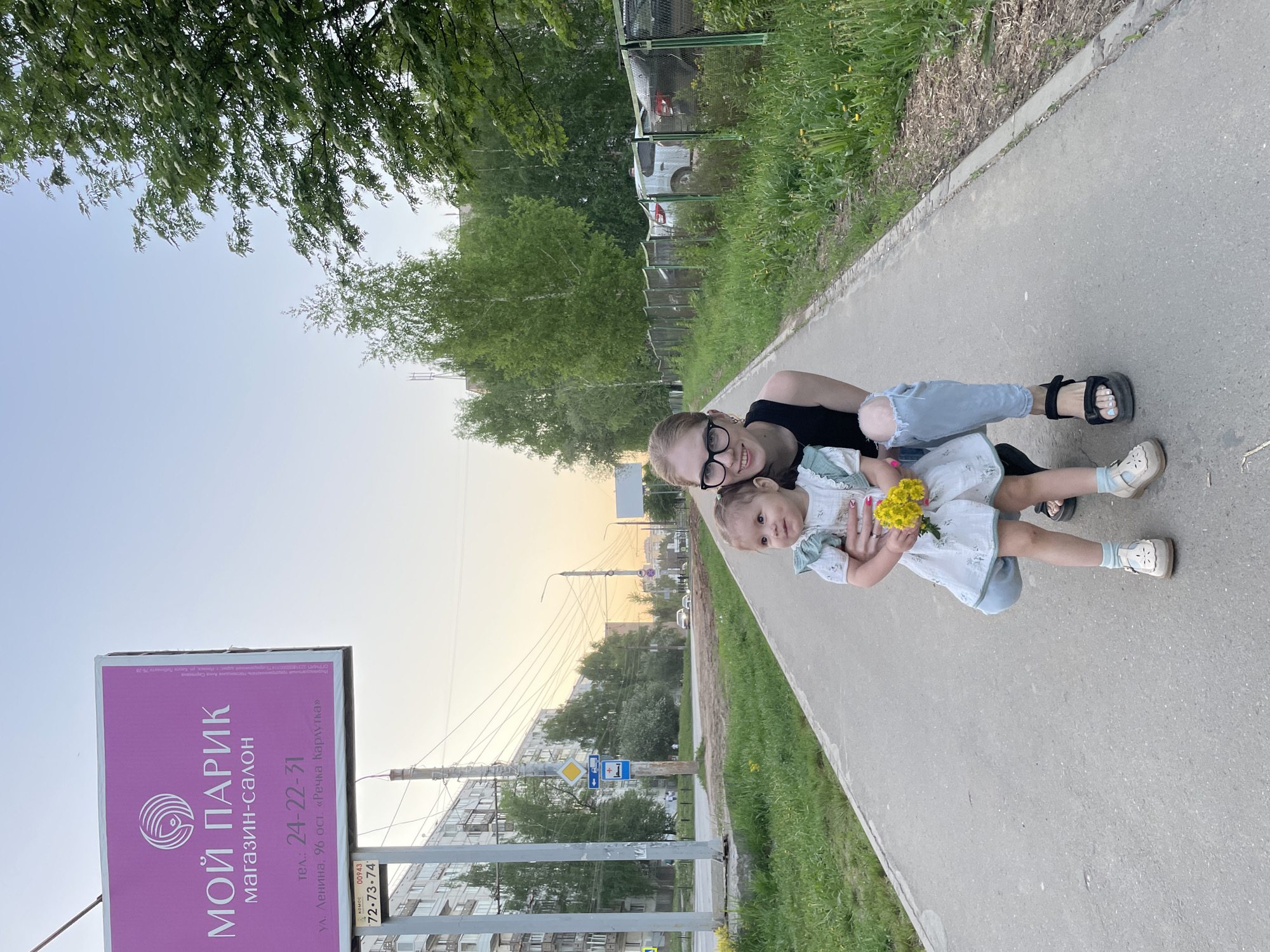 The image captures an outdoor scene at dusk, depicting an adult and a young child posing for a photograph. The adult lies on her back on a paved sidewalk, with the child lying on her, both looking towards the camera. The adult has blonde hair, wears black-rimmed glasses, a black top, and light blue ripped jeans, complemented by black sandals. She smiles broadly. The child, dressed in a light-colored dress with subtle patterns, holds a small bouquet of yellow flowers and wears light-colored shoes with socks, displaying a gentle expression.
To the left, a prominent pink signboard displays Russian text. It reads: "МОЙ ПАРИК магазин-салон" (MY WIG shop-salon), "тел.: 24-22-31", and "ул. Ленина, 96 ост. «Речка Карлутка»" (ul. Lenina, 96, stop "Rechka Karlutka"). Below this, additional text "комос 00943 72-73-74" is visible. This indicates a location on Lenina Street, near the "Karlutka River" stop, which is in Izhevsk, Russia.
The setting includes a strip of green grass bordering the sidewalk. Beyond the grass, various trees and bushes, along with wooden and chain-link fences, form a backdrop. Parked cars and distant residential buildings are visible through the foliage. Utility poles and overhead wires crisscross against the sky, which transitions from light yellow and orange near the horizon to a paler blue overhead, characteristic of late afternoon or early evening. A blue bus stop sign and a yellow diamond-shaped road sign are also present in the background. The scene conveys a calm and pleasant atmosphere.