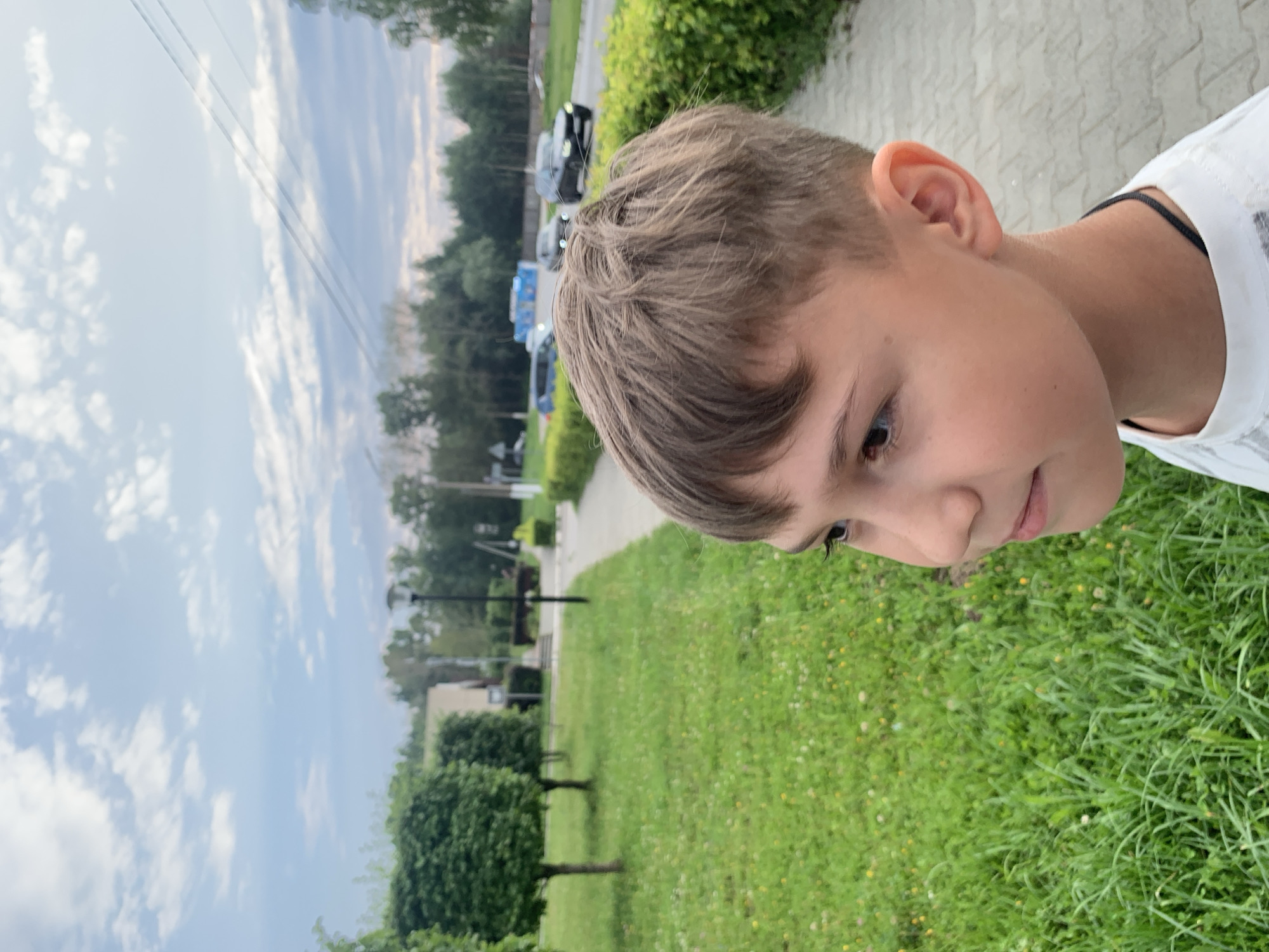 The image features a close-up, portrait-style shot of a young boy looking to his right, with his head tilted. He has light brown hair styled with some volume and appears to be wearing a white t-shirt with a dark neckline. The background suggests an outdoor setting in Chaikovsky, Russia.
To the boy's left, a cloudy sky dominates the upper portion of the frame, with what appear to be power lines stretching across. Below the sky, a street or pathway is visible, with several cars parked or driving in the distance, and a row of green bushes and trees. The ground is covered in green grass, dotted with small yellow flowers. The overall impression of the scene is casual and outdoors, likely during the daytime due to the bright sky. There is no visible text in the image.