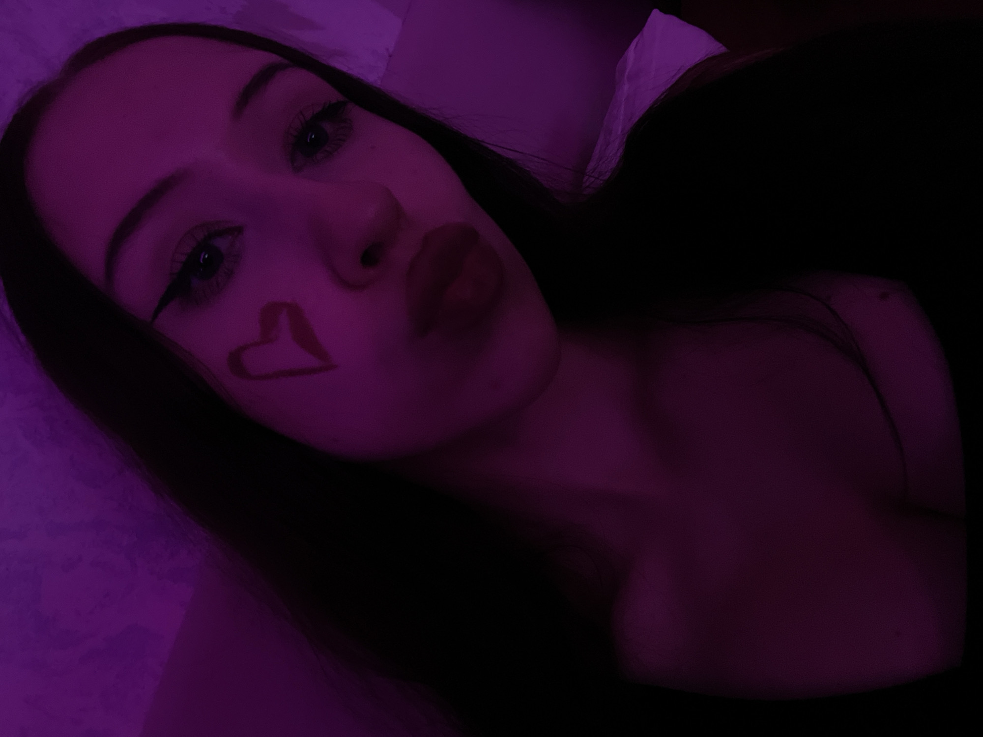 The image is a close-up selfie of a young woman with long, dark hair and a heart drawn on her left cheek. She is wearing dark clothing and has dramatic eyeliner. The scene is dimly lit with purple lighting, and the background appears to be a textured wall. There is no information in the image to suggest it is located in Votkinsk, Russia, or any specific urban environment.