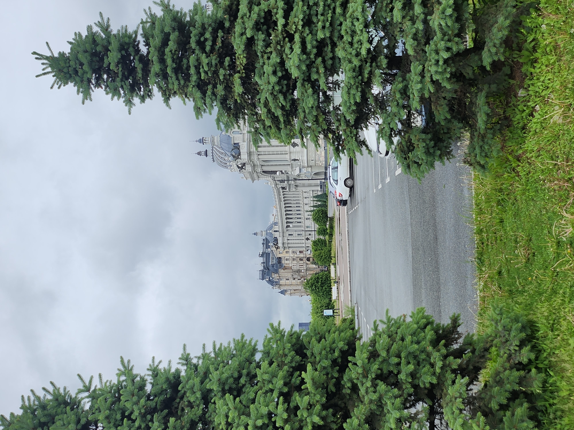 The image shows a view of the Palace of Farmers in Kazan, Russia, from a street lined with evergreen trees. The sky is overcast, and a white car is parked on the right side of the road. Green grass lines the edge of the road beside the trees.