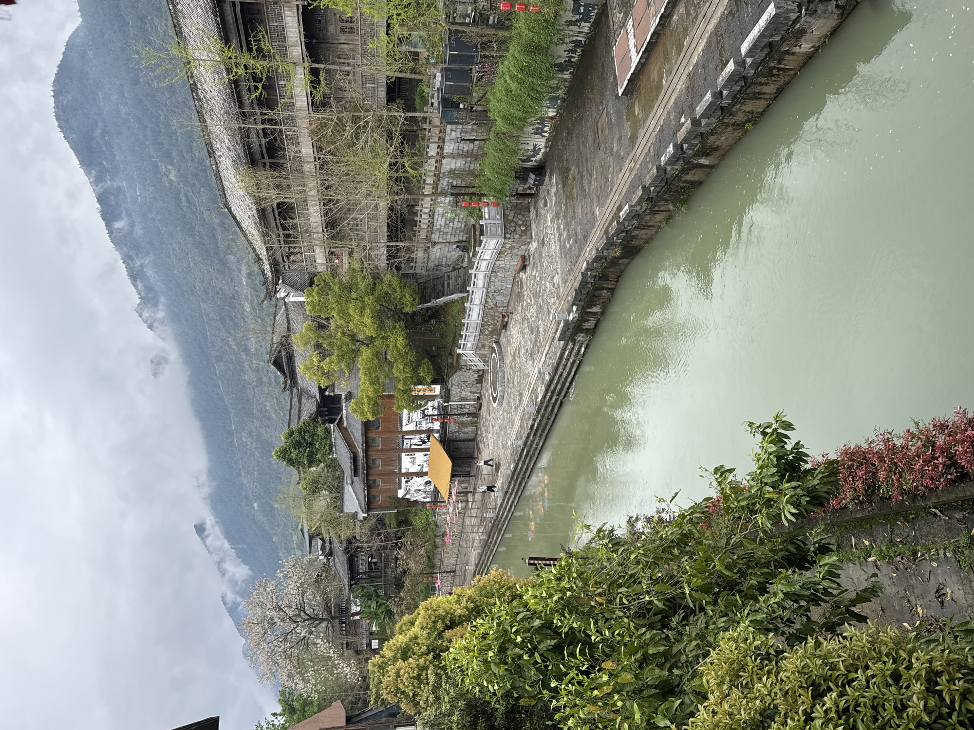 The image shows a view of Zhuoshui, China. A body of water runs along a stone embankment with buildings of traditional Chinese architecture visible on the other side. Mountains are in the background, partially obscured by clouds. Two people are walking on the stone embankment near a yellow awning. The foreground includes green foliage.
