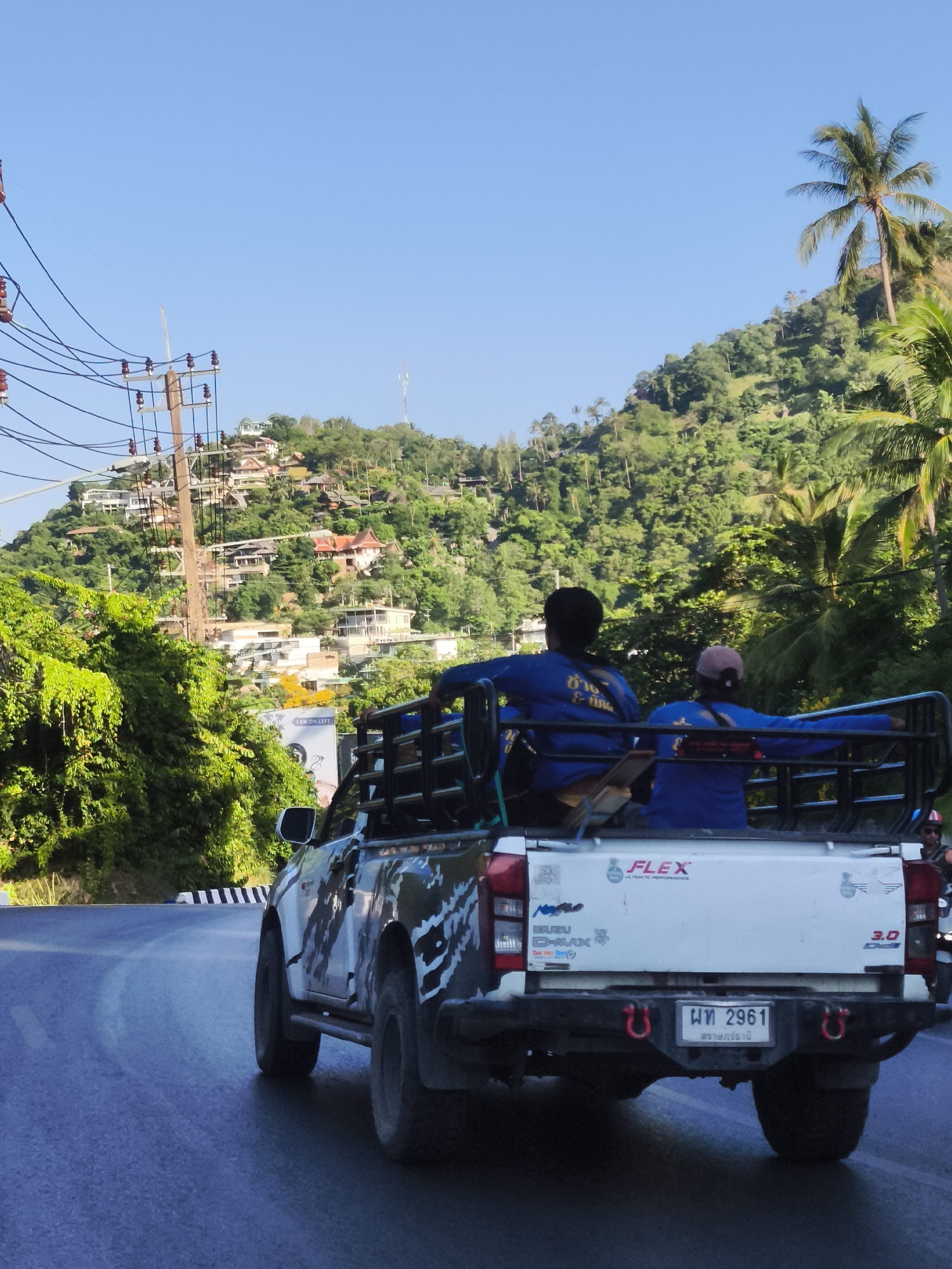 This image captures a pickup truck driving on a paved road in Ko Samui District, Thailand. The truck is white with a black and white camouflage-like design on its side. It is carrying two individuals in the bed of the truck, both wearing blue long-sleeved shirts. One person is facing away from the camera, while the other is looking slightly to the side. The truck has a metal rack in the back and is adorned with stickers, including one that reads "FLEX ULTIMATE PERFORMANCE." The background reveals a lush, green, mountainous landscape dotted with buildings, suggesting a residential area on a hillside. A clear blue sky stretches above. Power lines and utility poles are visible on the left side of the image. The road appears to be a two-lane road with white and black striped markings on the side. The overall impression is a scenic, possibly tropical, environment.