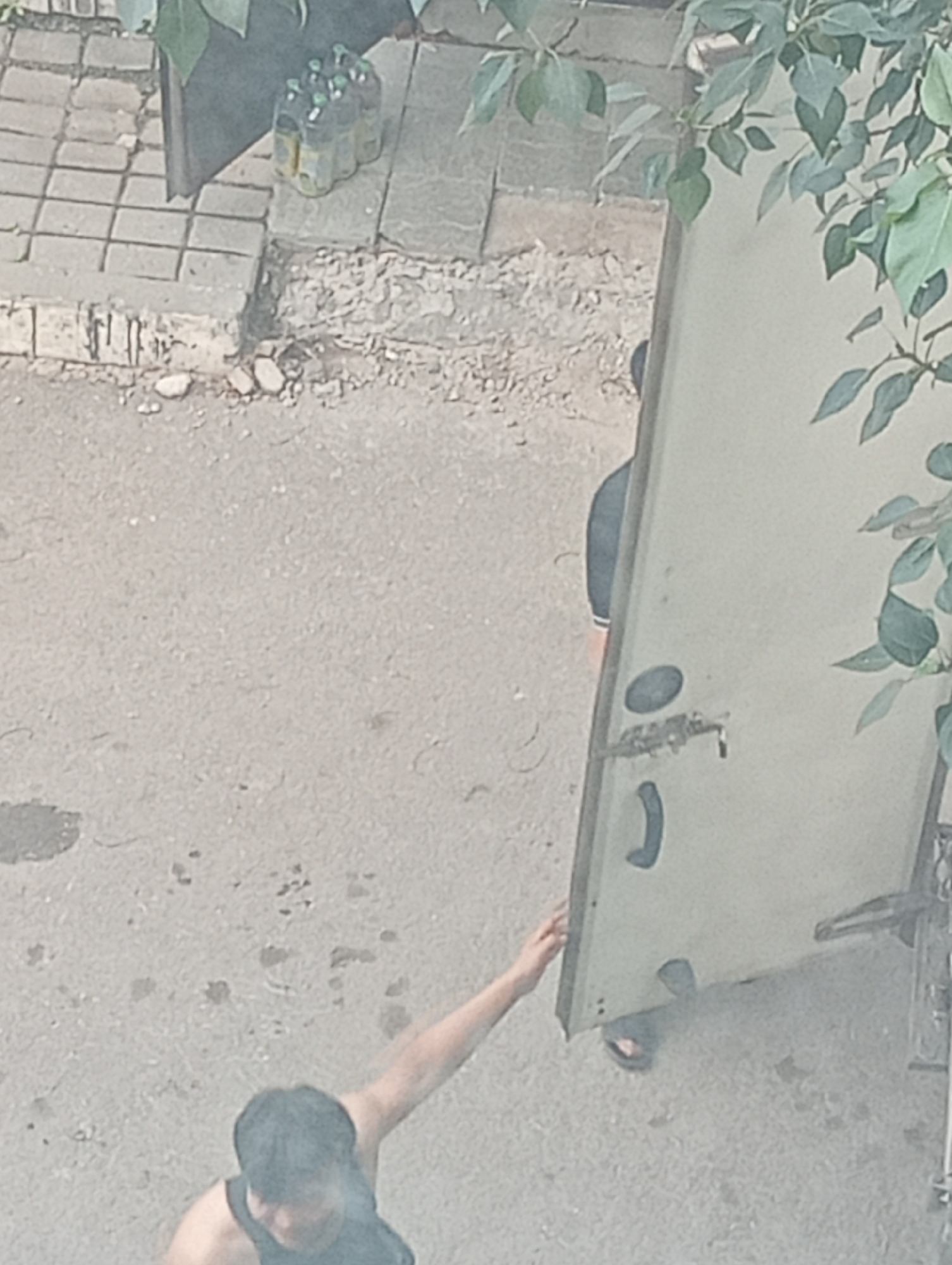 In an outdoor urban environment, an individual with dark hair and a dark tank top is seen from a high angle. They are extending their left arm towards a light-colored, rectangular object, possibly a door or panel, that is leaning against a wall. The object has a handle and what appears to be a lock mechanism. To the left of this object, a group of plastic bottles with green caps is visible on a tiled surface. The ground is a light-colored asphalt with some debris. Another individual, partially obscured, is visible behind the leaning object, wearing dark clothing. Lush green foliage hangs from above, casting shadows.