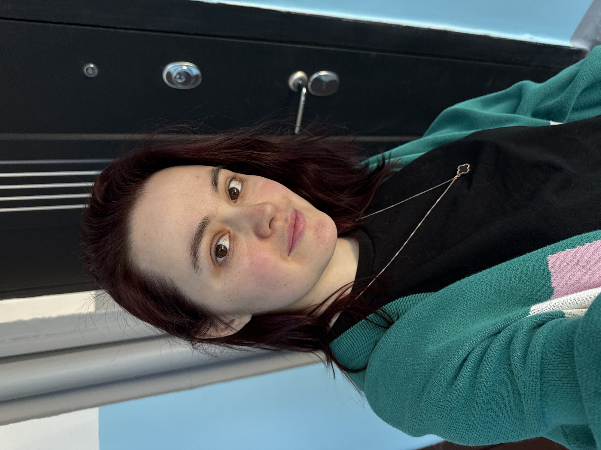 A photograph shows a woman with red-brown hair, fair skin, and brown eyes. She is wearing a black shirt and a green, pink, and white sweater. She also has a silver necklace with a four-leaf clover pendant. The background includes a dark-colored surface with circular fixtures and a lighter-colored wall with horizontal lines. It is not possible to determine the location in the city of Izhevsk, Russia.