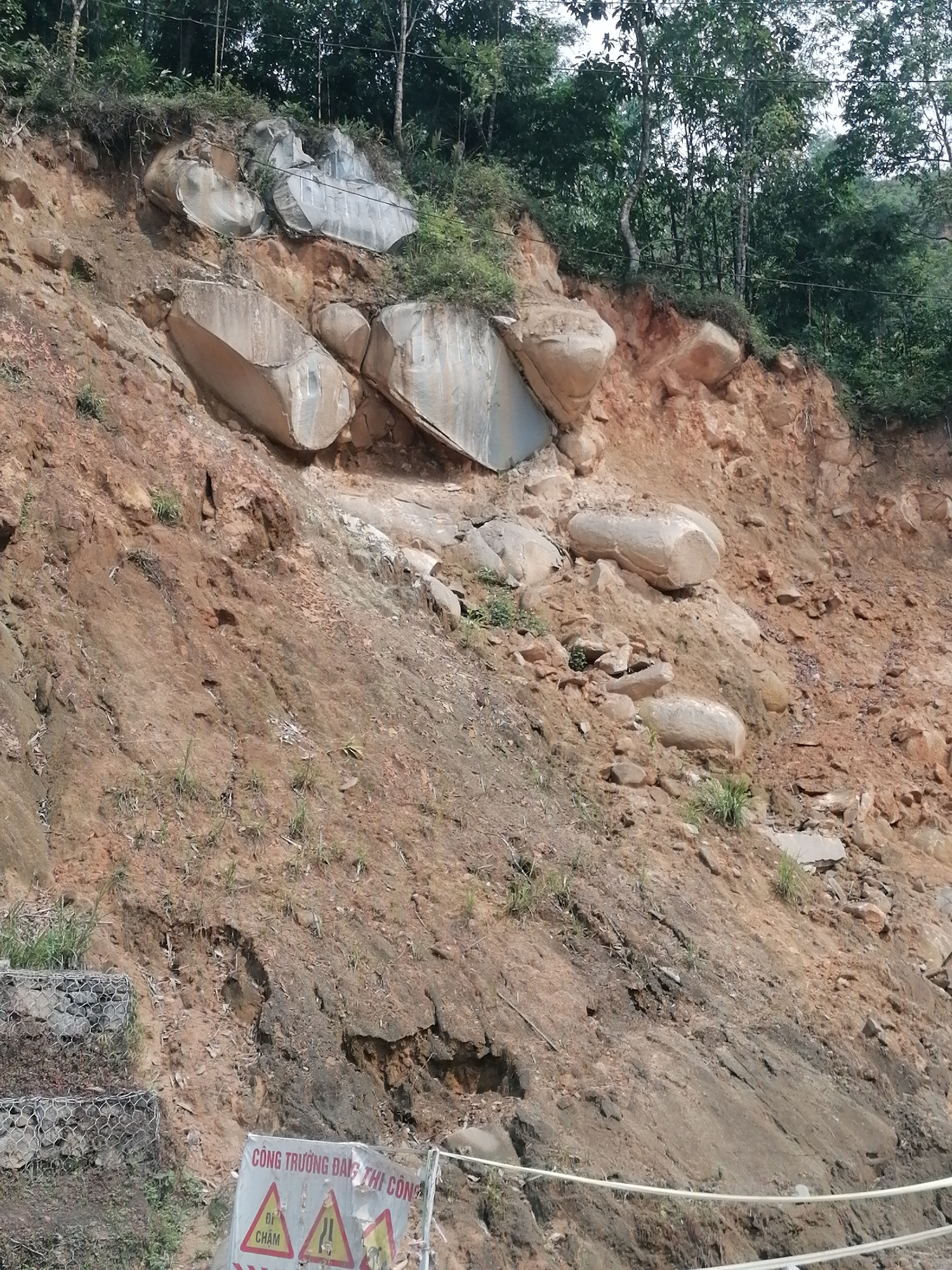 The image shows a hillside in Trung Chải, Vietnam, that has experienced a landslide. Large, roughly cylindrical rocks are embedded in the reddish-brown soil, some near the top of the hill. The top of the hillside is wooded. At the bottom, there is a sign indicating a construction zone. There is also wire mesh filled with rocks, likely for erosion control. A white rope acts as a barrier.
