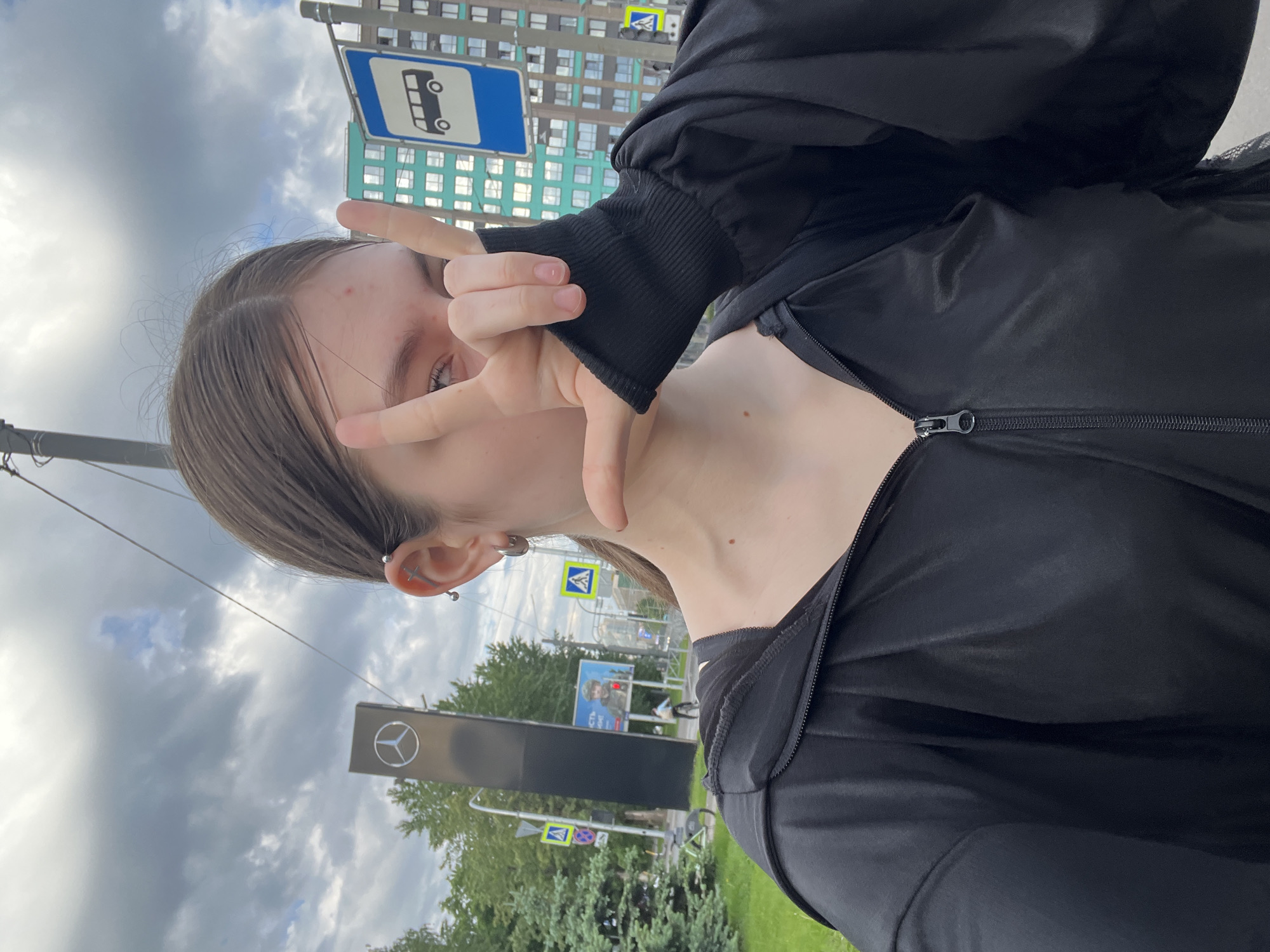 A vertical selfie-style image shows a young person taking a photo in an urban environment under an overcast sky. The person is positioned in the foreground, looking towards the camera, with their left hand raised to form a "peace" or "V" sign that partially covers their face. They have light brown hair parted down the middle and wear several ear piercings, including one shaped like a cross. They are dressed in a black zippered jacket, revealing a black top underneath. A small red blemish is visible on their forehead.
The background features a city street or sidewalk. A prominent blue rectangular sign with a white bus icon, indicating a bus stop, is visible near the top left. Behind this sign, a modern building with light blue-green panels and numerous windows can be seen. To the right, a large dark sign bearing the white Mercedes-Benz logo stands amidst green foliage. Other yellow triangular road signs, likely pedestrian crossing warnings, are also present in the mid-ground. The sky is predominantly cloudy. The overall impression is a casual outdoor shot in an urban setting. No clear text is readable apart from the Mercedes-Benz logo.