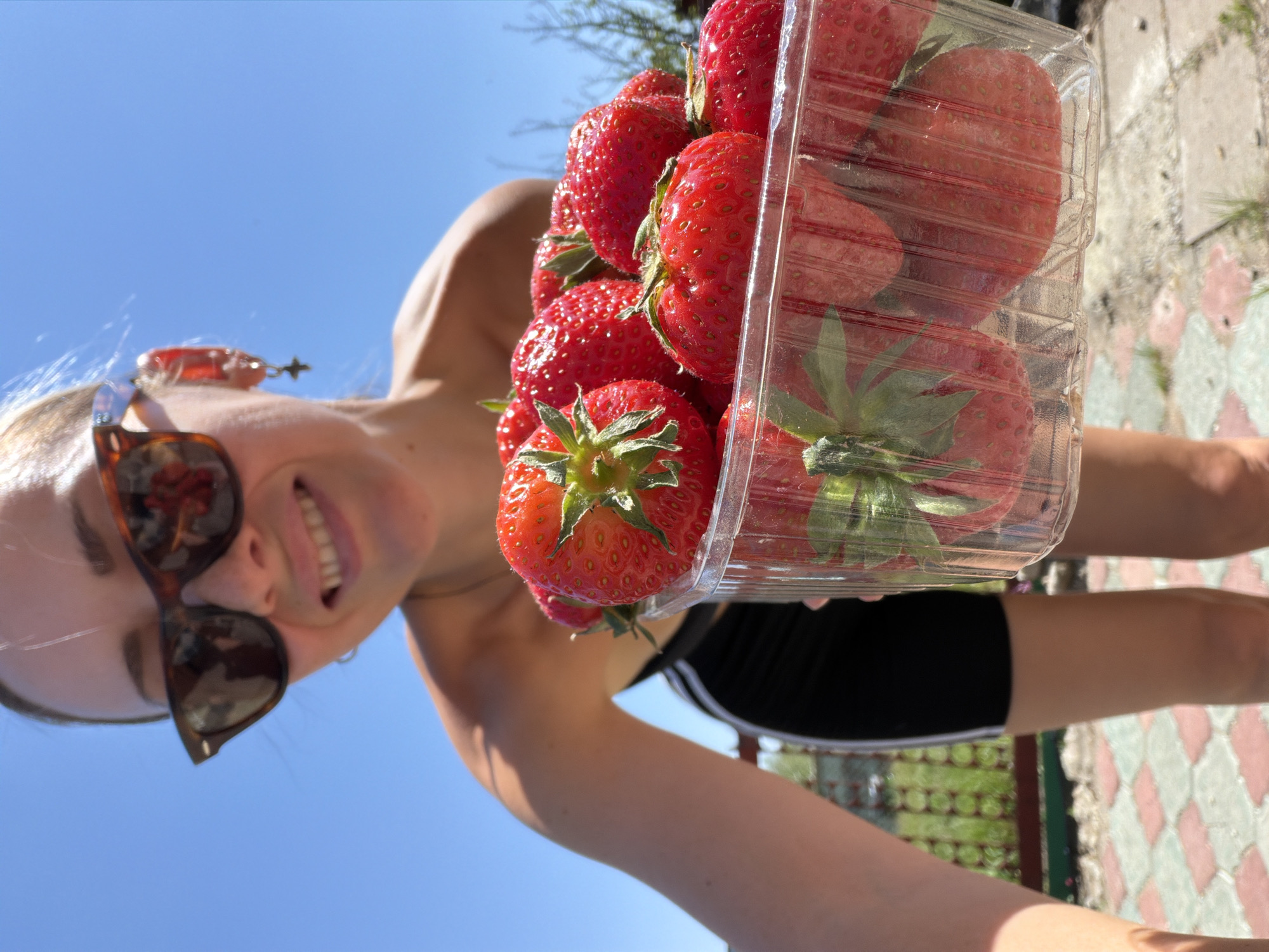 A young woman, wearing brown sunglasses and black shorts with white stripes, holds a clear plastic container filled with red strawberries in front of her. She is smiling at the camera. The background features a clear blue sky, a glimpse of green foliage, and a stone-paved ground. The location is identified as Kamyshin, Russia.