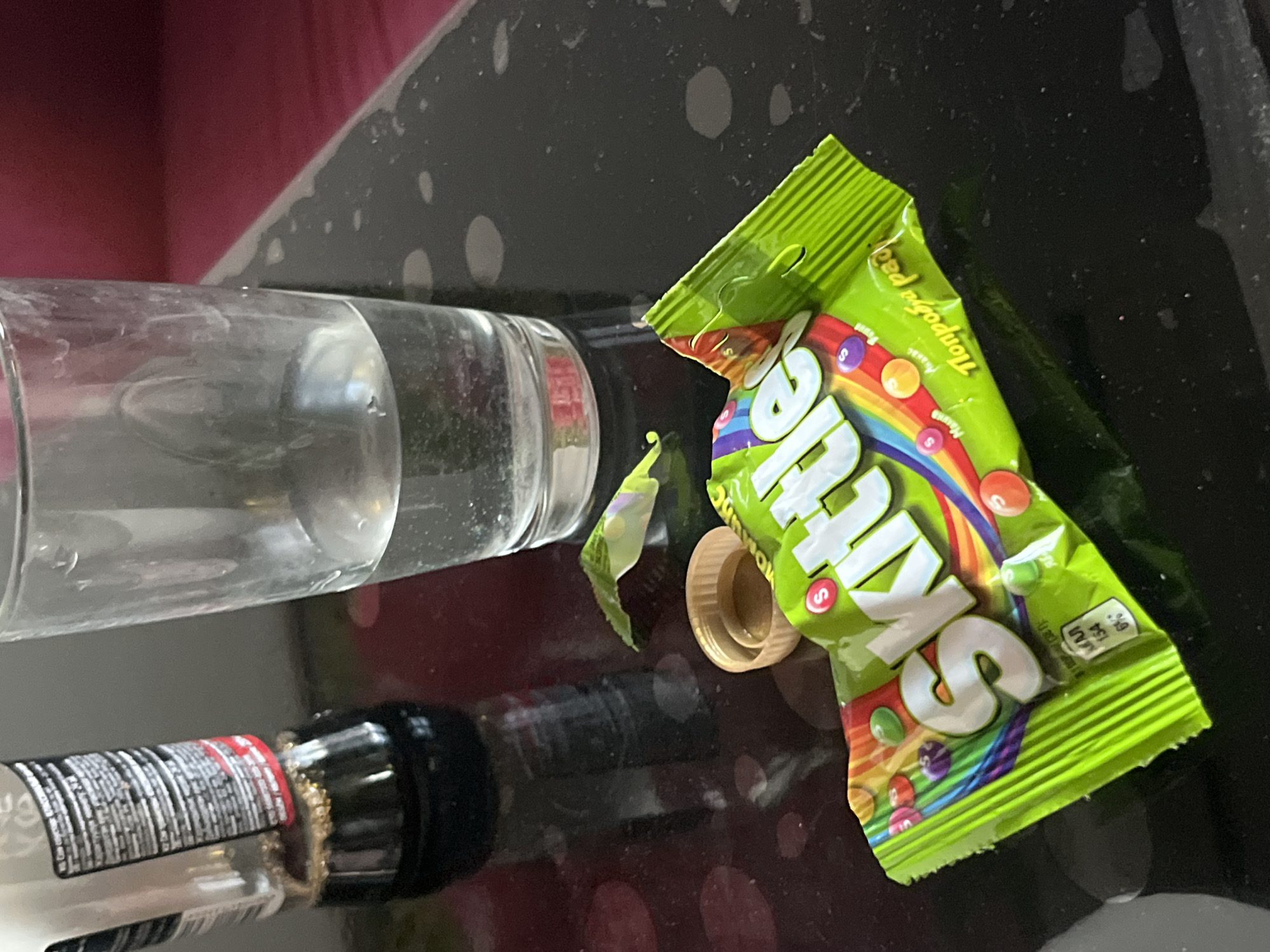 The image depicts a close-up of a countertop with various items. A partially filled clear glass, a black bottle with white text, and an opened green Skittles candy package are visible. The Skittles package is lying on its side with its tan plastic cap beside it. The background includes a dark, possibly black countertop with light colored, round shapes on it, and a section of a red surface. There is no evidence of a specific city or location. Moscow, Russia cannot be confirmed based on the visual information.