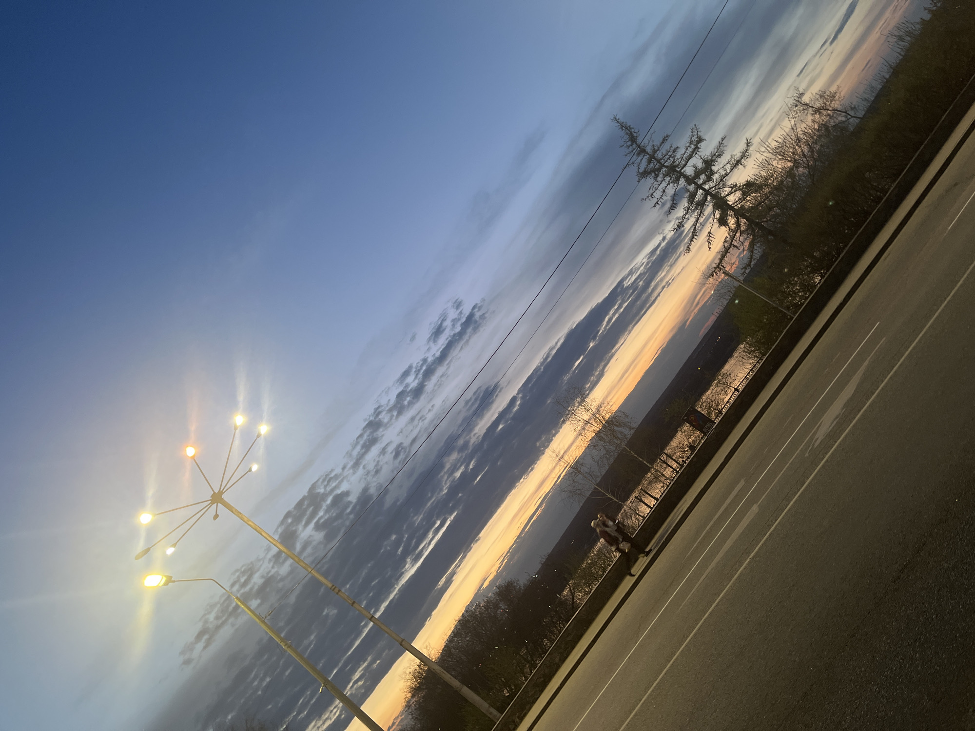 The image shows a view of the Kama River embankment at sunset in Perm, Russia. The photo is taken at an angle, revealing the asphalt road with white lines running parallel to the embankment.
The sky exhibits a gradation of colors, transitioning from a deep blue overhead to a lighter hue closer to the horizon, where the setting sun casts an orange and yellow glow. Stratus clouds are present in the sky.
A large, modern street lamp with multiple spherical lights stands on the left side of the frame.
On the other side of the road is a metal barrier that is installed along the embankment. Behind the barrier, trees and vegetation are visible. The Kama River is visible in the middle ground, reflecting the light of the setting sun. Two people are standing in the distance along the embankment.
Electrical wires stretch across the sky.