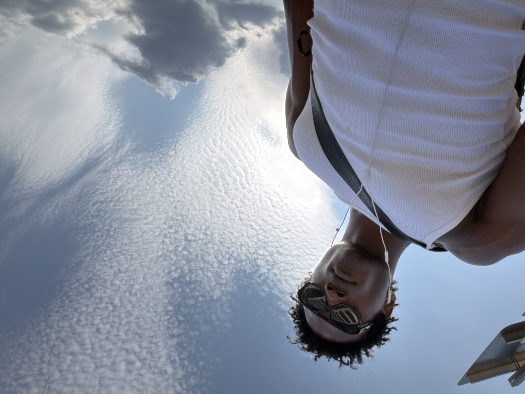 A low-angle shot captures a dark-skinned person wearing a white ribbed tank top, black sunglasses, and white earbuds, looking upwards towards a sky with scattered clouds. The person has dark, curly hair and a strap across their chest. To the left of the frame, a modern, geometric sculpture is visible against the blue sky. The background consists of a dynamic sky with both fluffy white clouds and some darker, more ominous clouds. The overall lighting suggests it is daytime. The location appears to be Liberty State Park in Jersey City, USA, given the presence of the sculpture which resembles the "The New Colossus" monument by the artist Pi Li, and the iconic view of the New York City skyline in the distance, though the skyline is not visible in this particular frame.