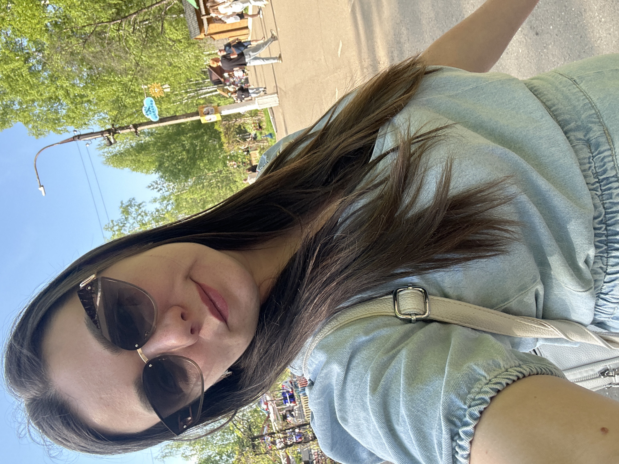 A selfie taken by a woman with long brown hair wearing sunglasses and a light blue denim outfit with a white purse strap. The background appears to be an outdoor scene in the city of Chaikovsky, Russia, with trees, a street, and people walking around. A light post is visible on the left side of the frame. In the distance, there's a possible fair ride visible.