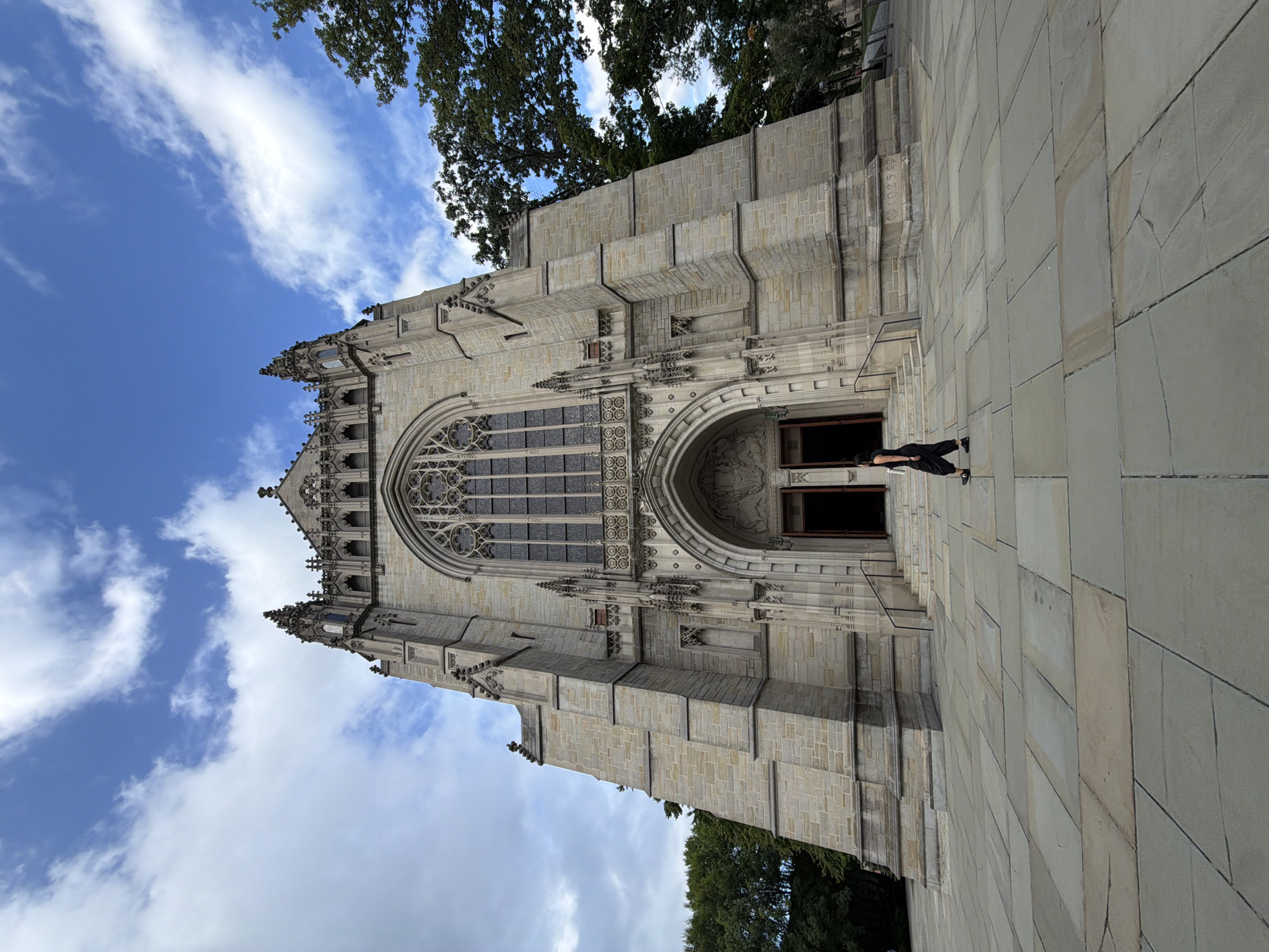 A low-angle, full-shot photograph captures the exterior of the Princeton University Chapel in Princeton, New Jersey. The Gothic Revival-style chapel, constructed of light-colored stone, dominates the frame, its ornate facade featuring a large rose window and a grand arched entrance. Two dark doorways are visible within the entrance arch. The building's spires and intricate stonework rise towards a blue sky scattered with white clouds. In front of the chapel, a woman dressed in a black jumpsuit walks across a wide, paved plaza composed of large, rectangular stone tiles. Trees with lush green foliage flank the right side of the chapel. The overall lighting suggests daytime.