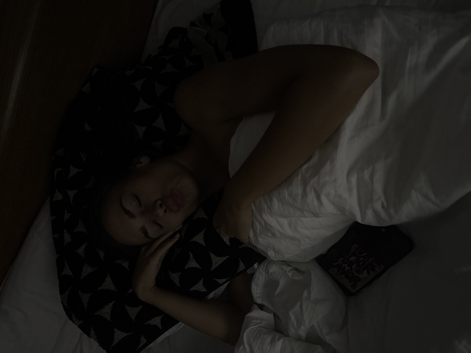 A woman lies asleep on a bed, face up on a dark patterned pillow. Her left arm rests near her face. She is partially covered by white sheets, with a dark object visible beneath. The scene is dimly lit, suggesting nighttime. The bed is made with white linens, and a wooden surface is visible at the edge of the frame.