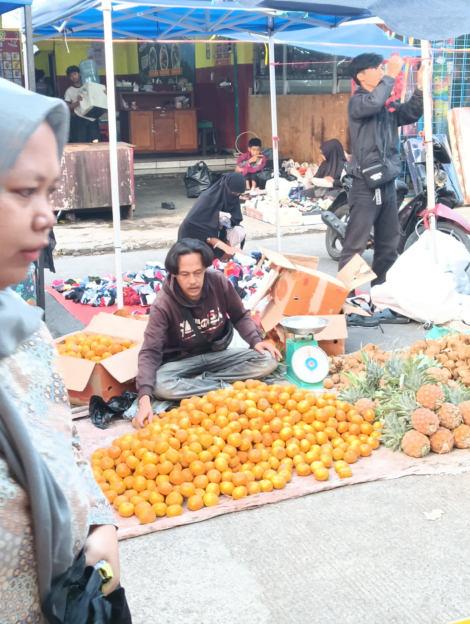 The image captures a scene at an outdoor market in Katapang, Indonesia. In the foreground, a large pile of bright orange fruits, likely tangerines or mandarins, is displayed on a tarp. To the right of the fruit, a cluster of pineapples is visible. A man wearing a brown hooded sweatshirt and blue jeans sits on the ground next to the fruit, with his hands near the oranges. A green mechanical scale is placed beside the oranges and pineapples. In the mid-ground, various items of clothing are spread out on a red tarp. Several individuals are visible in the background, including a young boy sitting and looking at something in his hands, a woman in a black hijab, and a person wearing a black jacket and black pants, who appears to be adjusting something. Behind them, a blue tent structure is set up, and a motorcycle is parked. The ground is paved with concrete. The overall atmosphere suggests a busy marketplace environment.